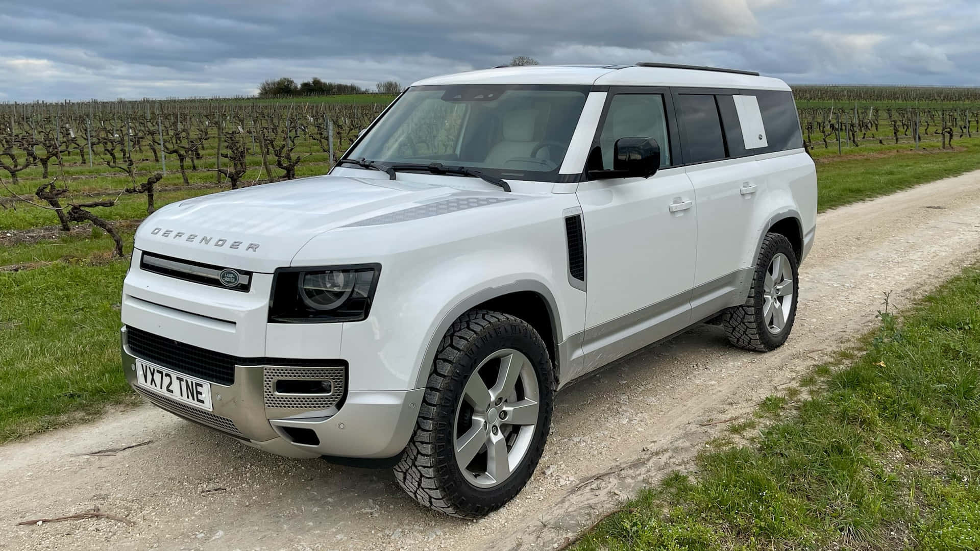 Land Rover Defender 桌布 壁紙 — 一輛白色 Land Rover Defender 停泊在碎石路上，旁邊是休眠中的葡萄藤行，天空多雲。