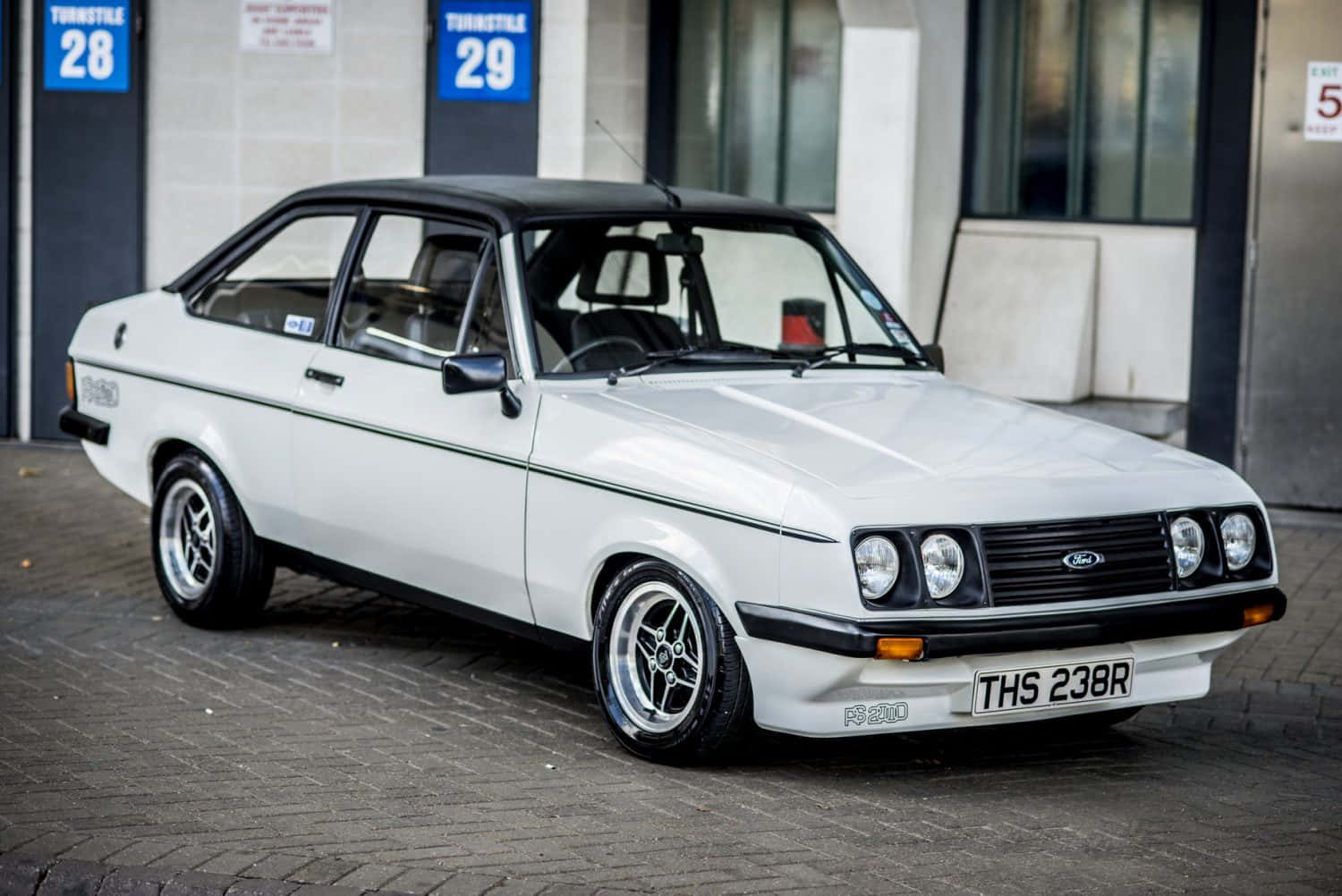Ford Escort 桌布 壁紙 — 一輛純白色的福特 Escort RS1600，帶有黑色飾條和賽車輪圈，停在磚砌的車庫前院。