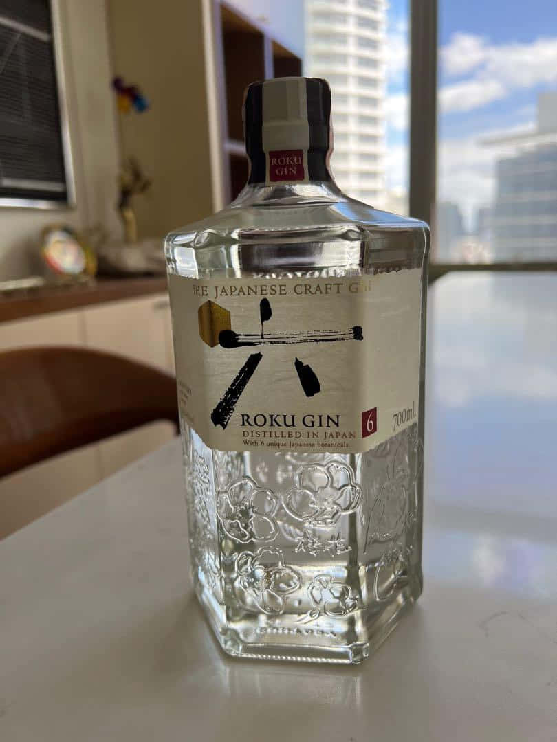  壁紙 — 一瓶清澈的Roku Gin，瓶身有花卉雕刻，放在白色檯面上，透過後面的窗戶可以看到城市天際線。
