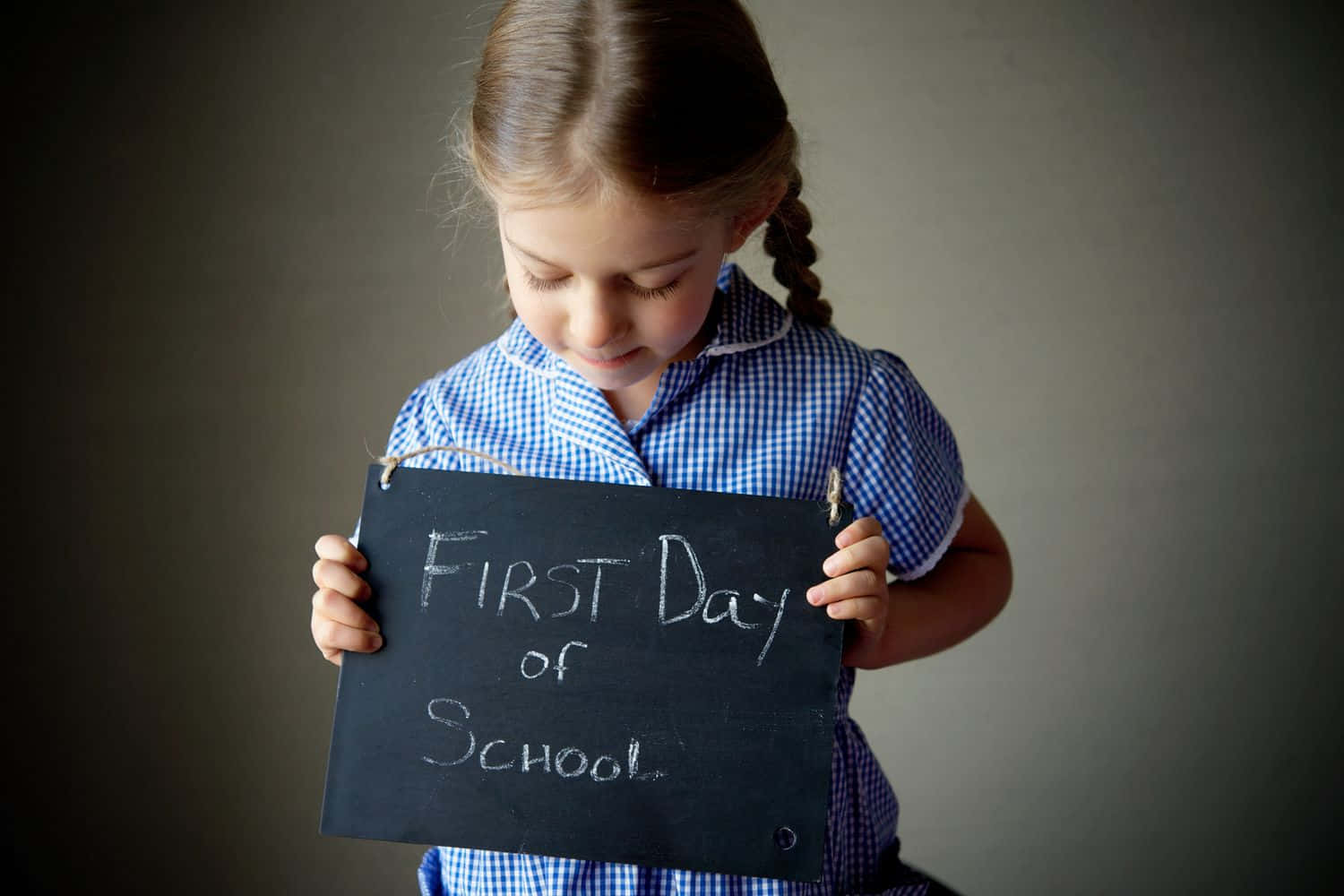  壁紙 — 一個穿著藍色格仔校服的年輕女孩，專注地拿著一塊寫著「First Day of School」的黑板標語牌。
