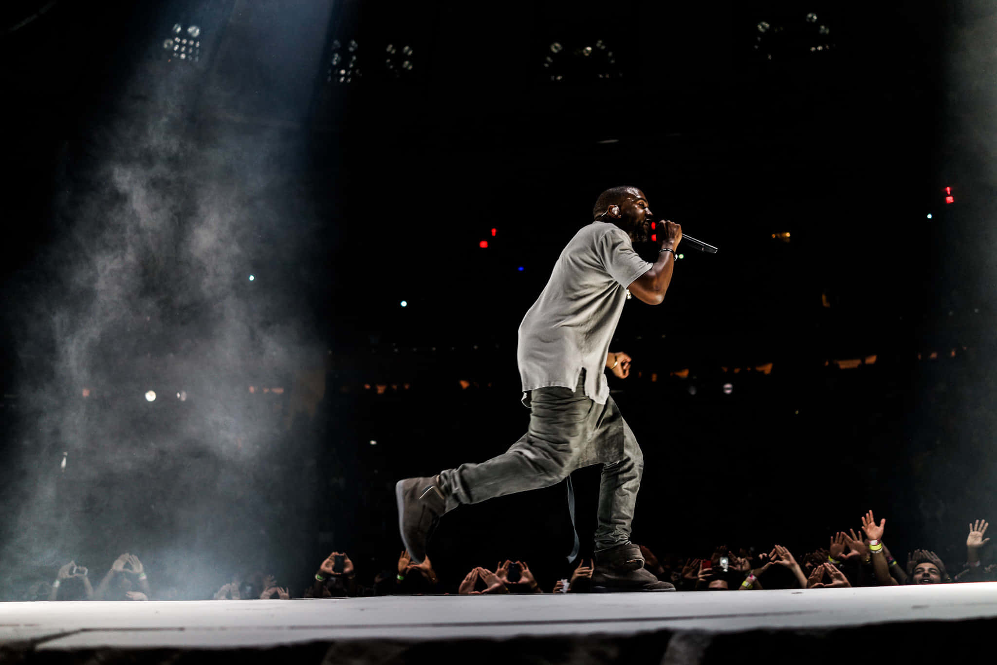 「Yeezus」— 喬治·威斯特 壁紙 — 表演者在明亮的聚光燈下，在煙霧瀰漫的舞台上動態地大步走過，下方熱情的觀眾伸手向上。