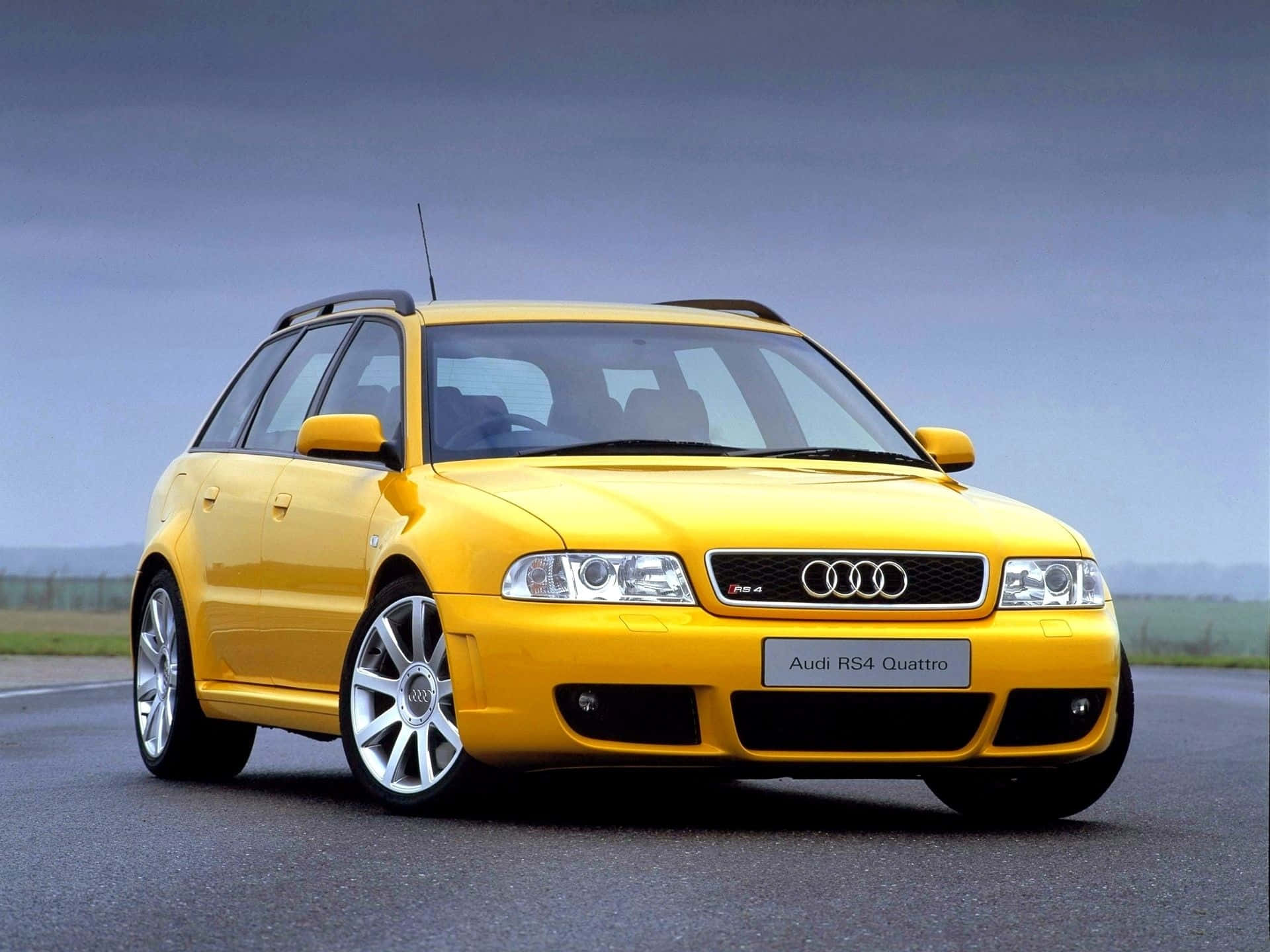 Audi Rs4 桌布 壁紙 — 一輛鮮黃色的奧迪RS4 Quattro旅行車停泊在瀝青路上，天空多雲。