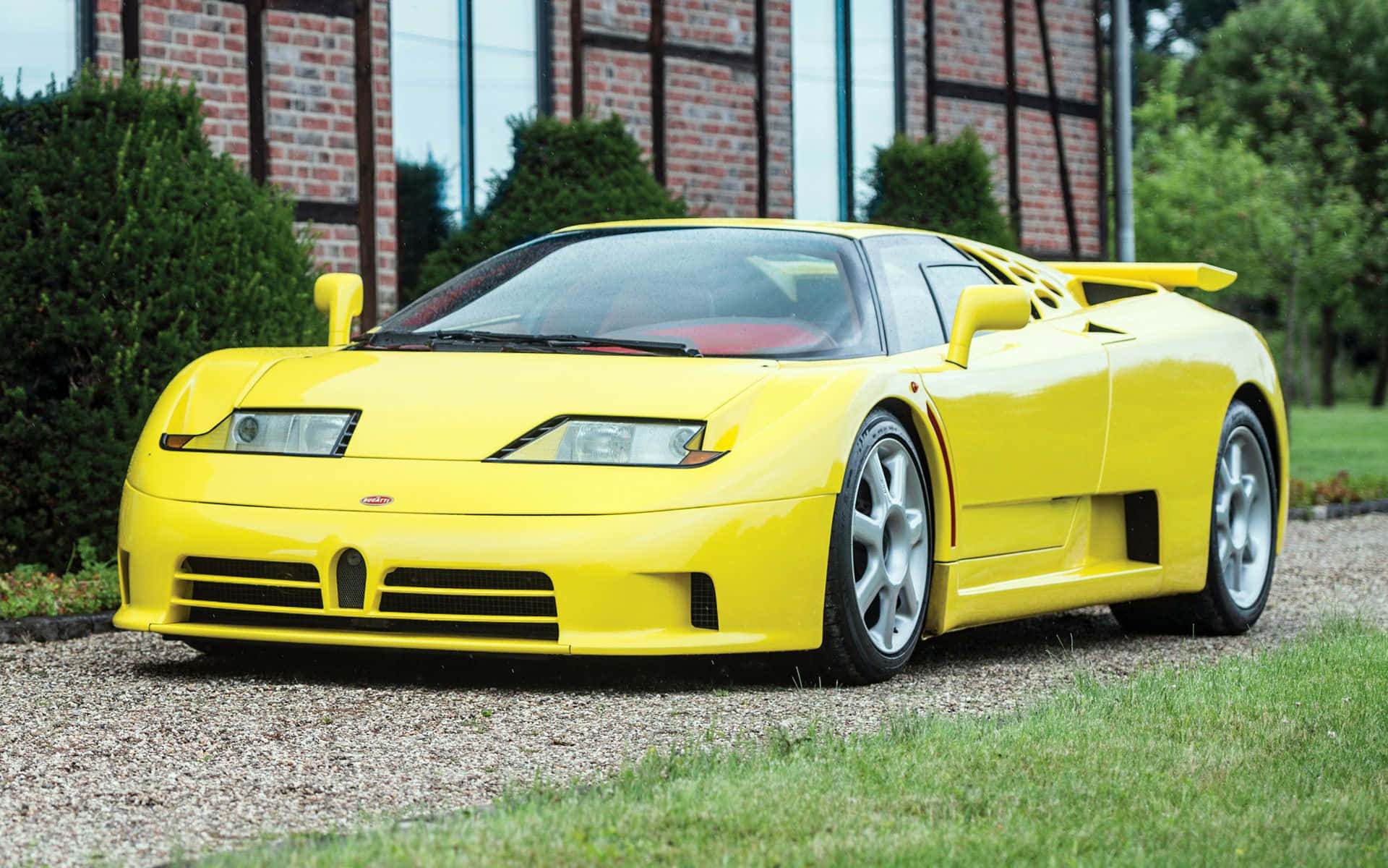 Bugatti Eb110 桌布 壁紙 — 一輛鮮黃色的Bugatti EB110超級跑車停泊在磚砌建築前的碎石地上，周圍有綠植。