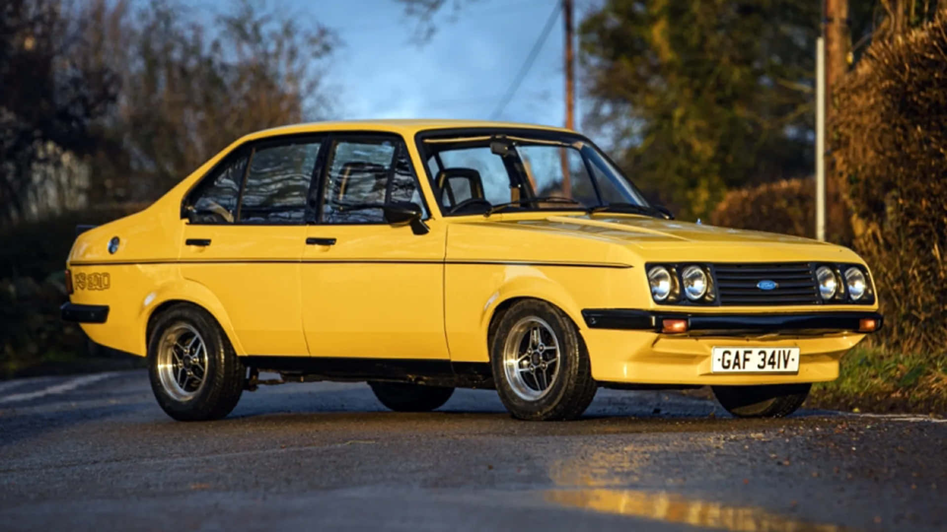 Ford Escort 桌布 壁紙 — 一輛鮮黃色經典福特Escort拉力賽車在黃金時刻停泊在碎石路上，背景有樹木。
