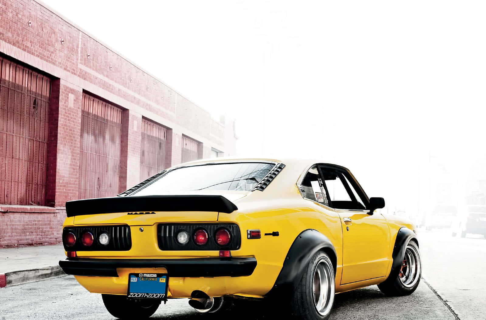 Mazda Rx-3 桌布 壁紙 — 一輛鮮黃色經典萬事得RX-7跑車，帶黑色點綴，停在磚砌倉庫旁，在明亮日光下。