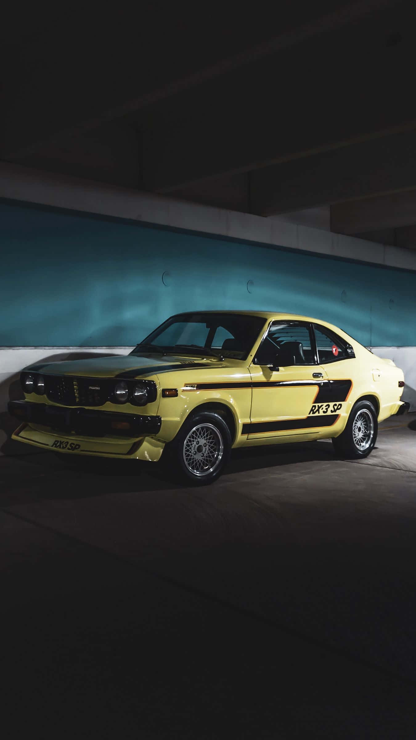 Mazda Rx-3 桌布 壁紙 — 一輛充滿活力的黃色經典RX-3 SP跑車在深色混凝土天橋下，帶有藍綠色牆壁點綴，被戲劇性地照亮。