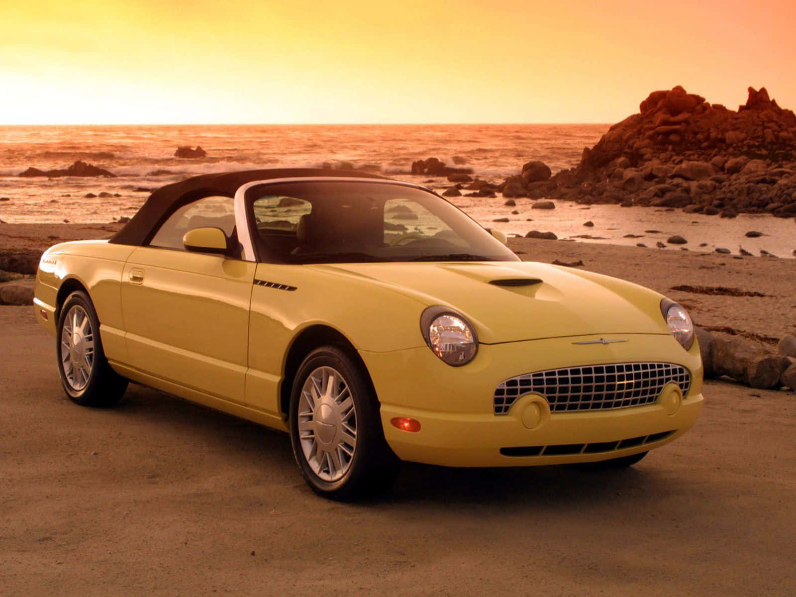 Ford Thunderbird 桌布 壁紙 — 一輛黃色開篷跑車停泊在岩石海灘上，夕陽西下，溫暖的橙色光線反射在海面上。