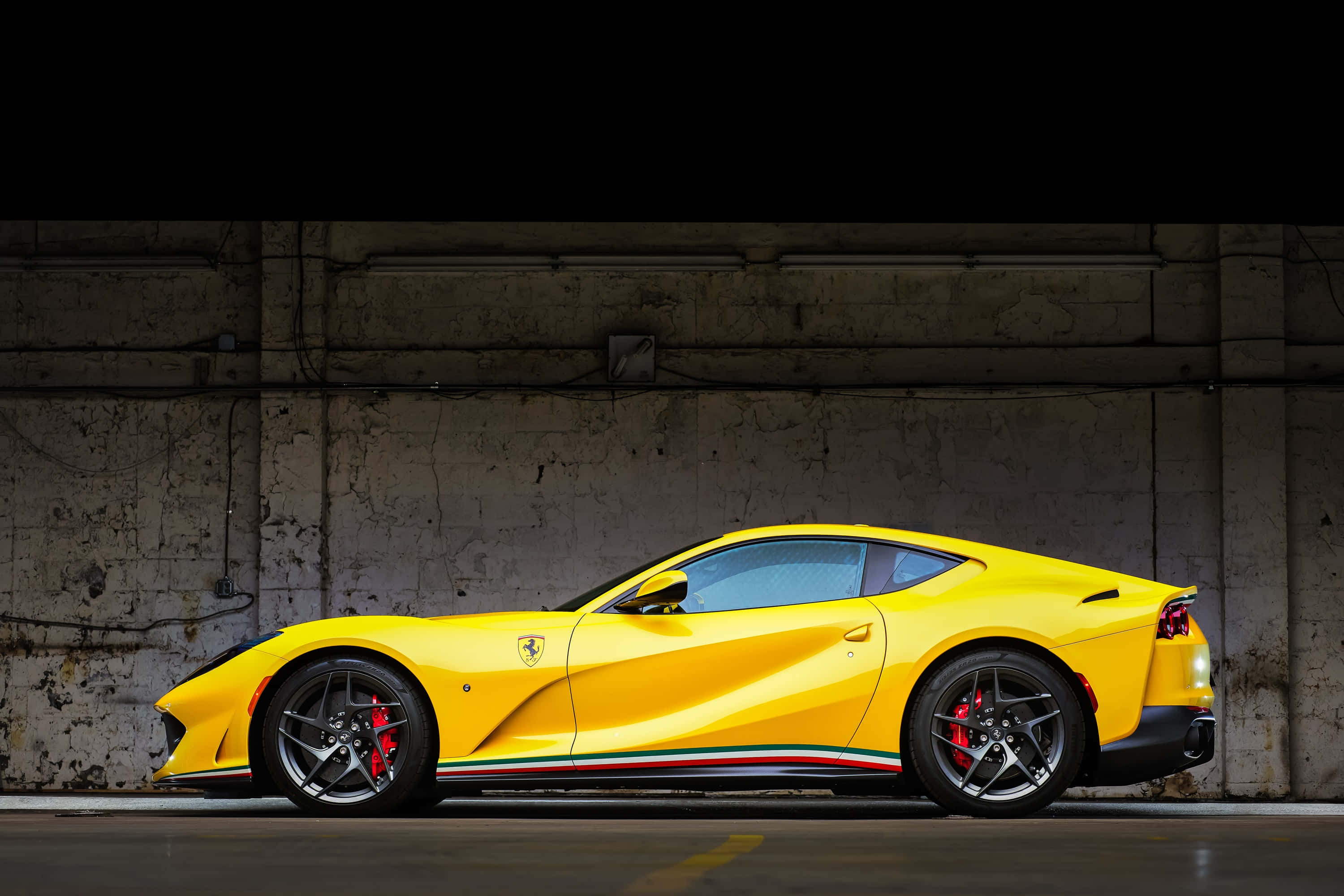 Ferrari 812 Superfast 桌布 壁紙 — 一輛鮮黃色法拉利跑車，配備紅色制動卡鉗和三色條紋，停放在光線昏暗的混凝土車庫中。