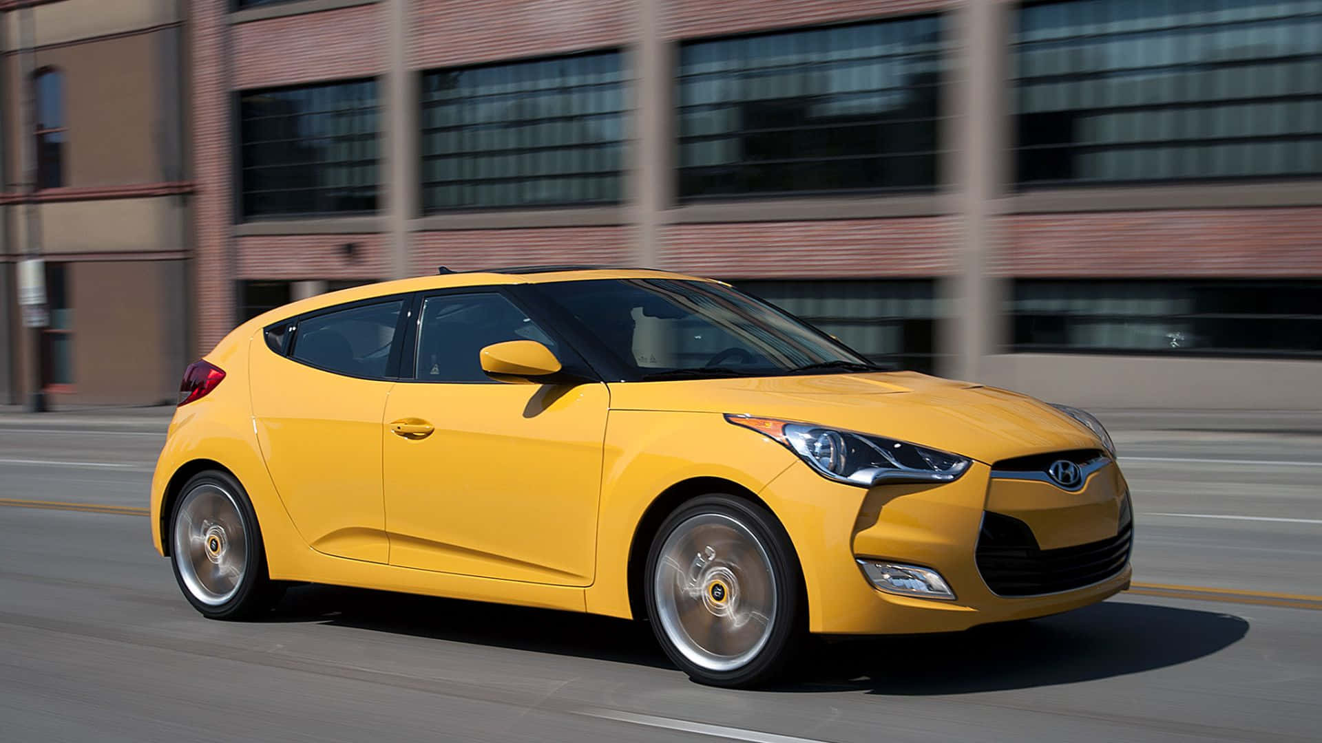 Hyundai Veloster 桌布 壁紙 — 一輛充滿活力的黃色現代Veloster在磚砌建築背景下，以動態模糊效果在城巿街道上飛馳。