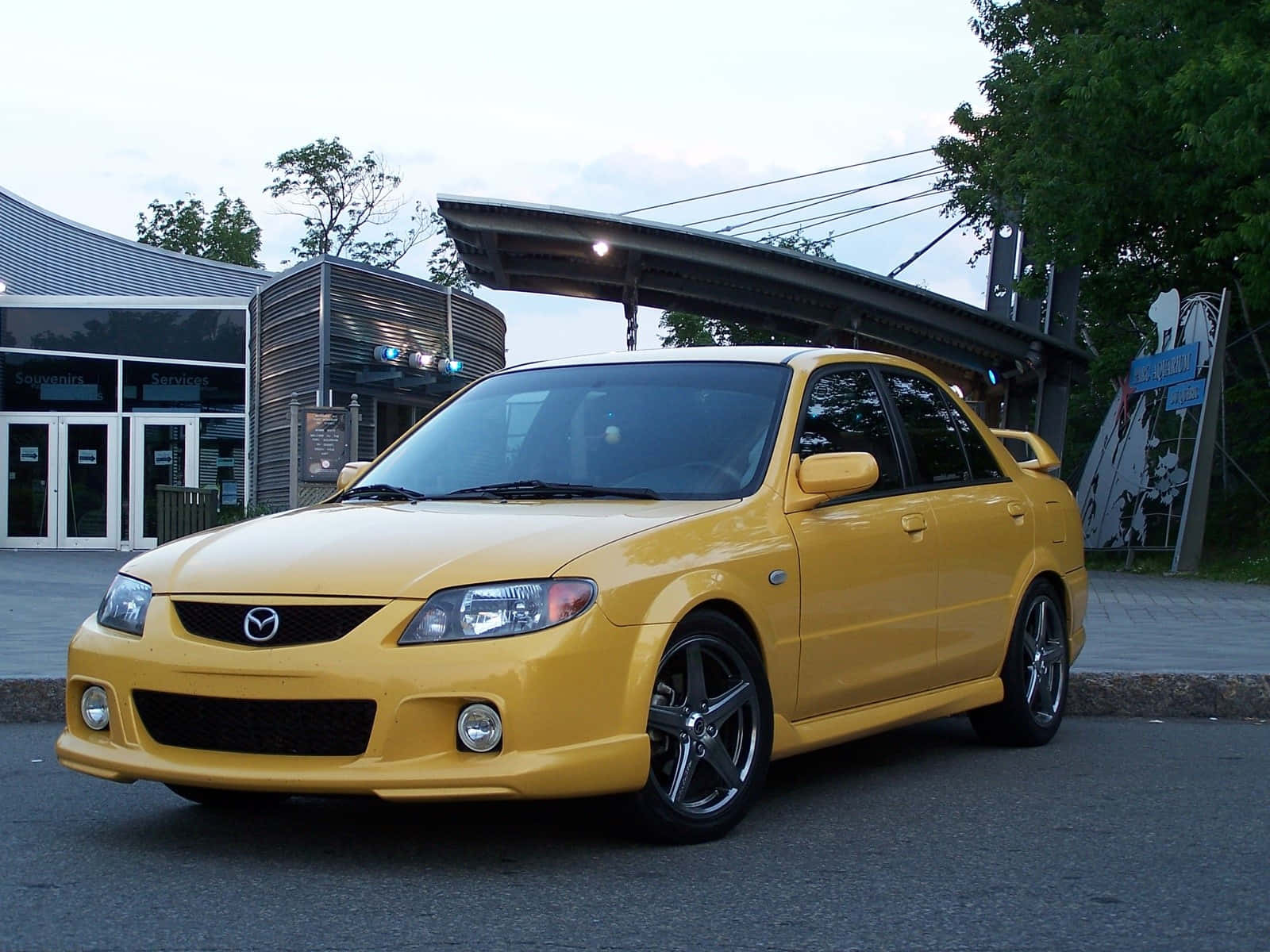 Mazda Protege 桌布 壁紙 — 一輛鮮黃色嘅Mazda 3房車，配黑色輪圈，喺黃昏時停泊喺現代建築附近嘅瀝青路上。