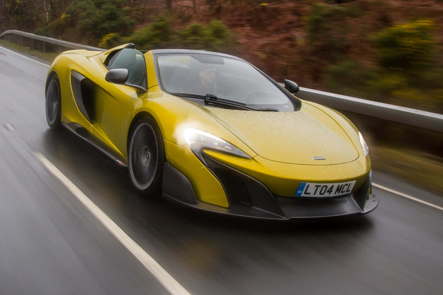 Mclaren 675lt Spider 桌布 壁紙 — 一輛充滿活力的黃色McLaren跑車在潮濕的道路上行駛，背景是模糊的綠色植物。