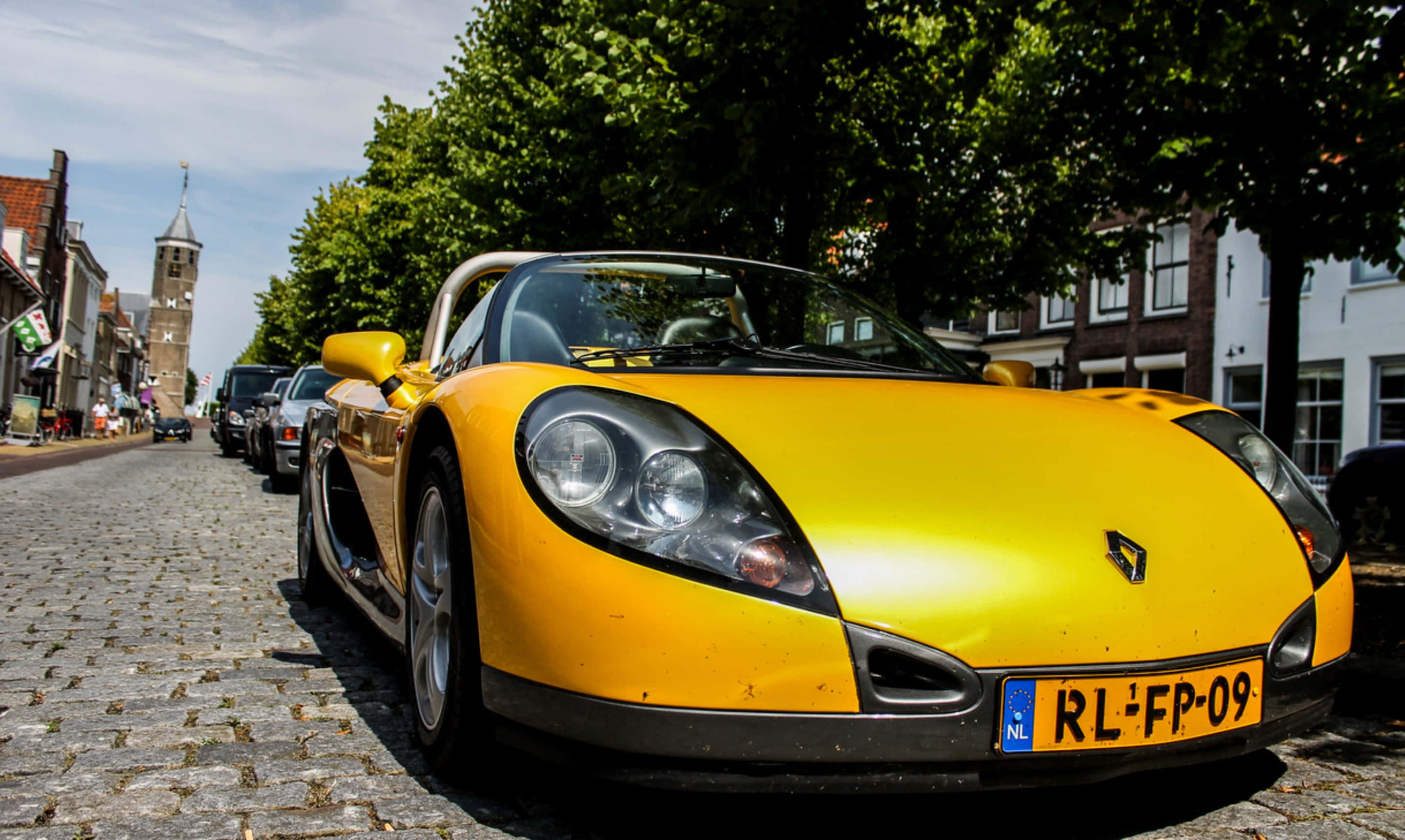 Renault Sport Spider 桌布 壁紙 — 一輛亮黃色跑車停泊在鵝卵石街道上，街道兩旁種滿樹木和歷史建築，天空晴朗。