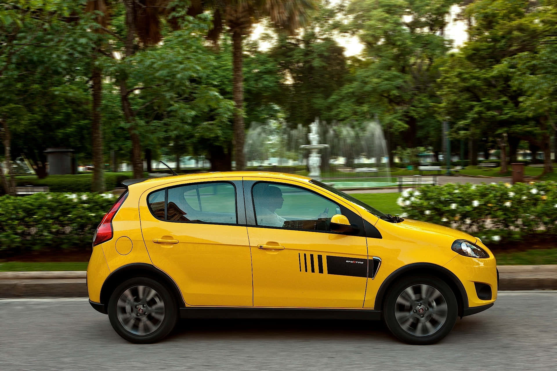 Fiat Palio 桌布 壁紙 — 一輛亮黃色的士高速駛過一個綠意盎然的公園，背景有噴泉。