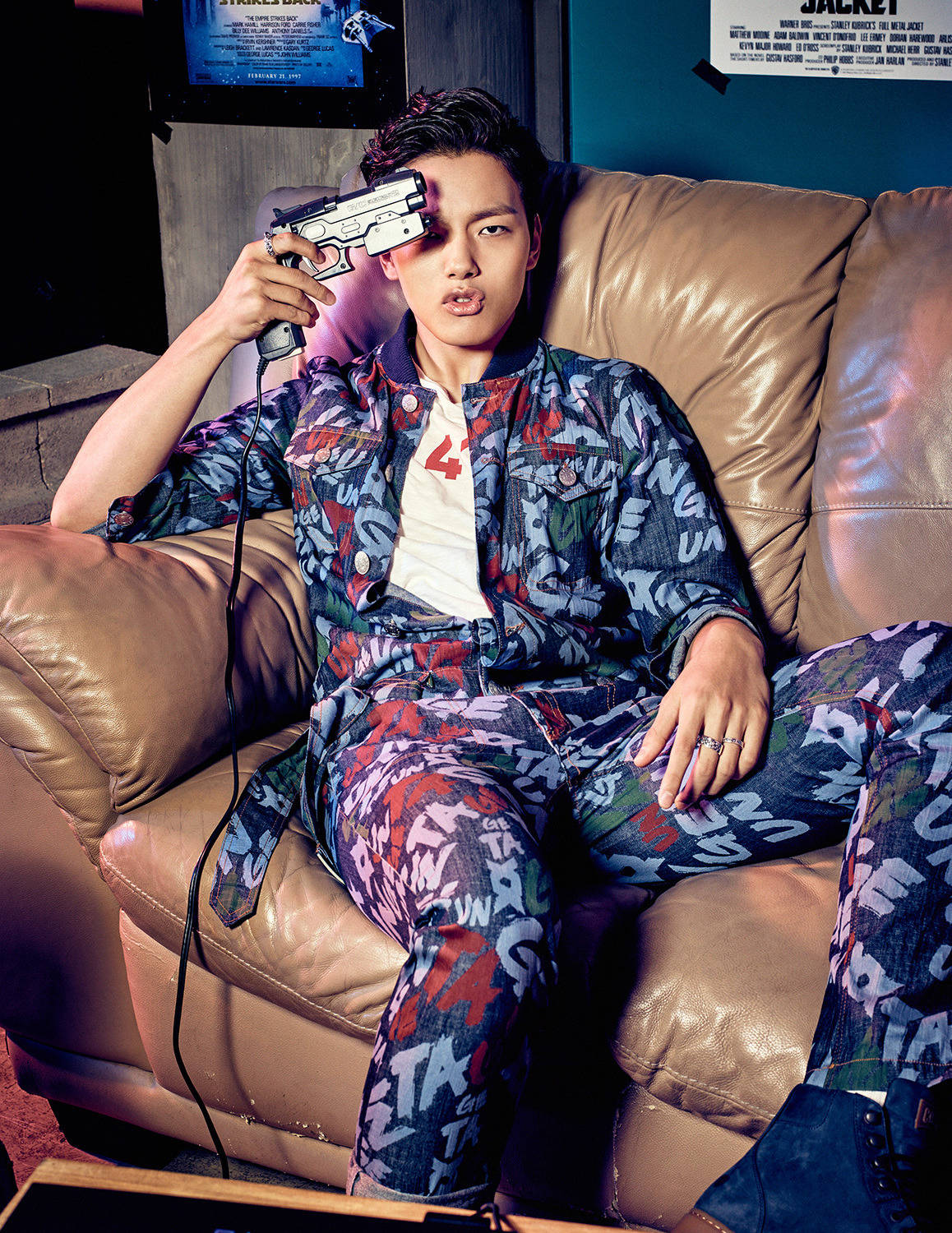 Yeo Jin Goo 為《Elle》拍攝時尚寫真 壁紙 — 一位身穿塗鴉圖案服裝的年輕男子懶洋洋地躺在皮沙發上，手持有線玩具槍，表情俏皮。