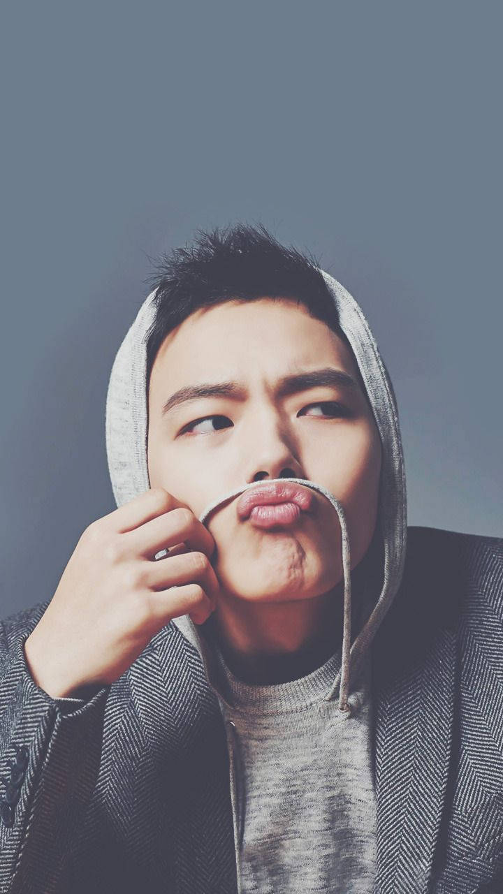 Yeo Jin Goo 嘴脣微翹展現迷人魅力 壁紙 — 一名身穿灰色連帽衛衣的年輕男子，在柔和的藍色背景前，用抽繩俏皮地拉著自己的嘴唇。
