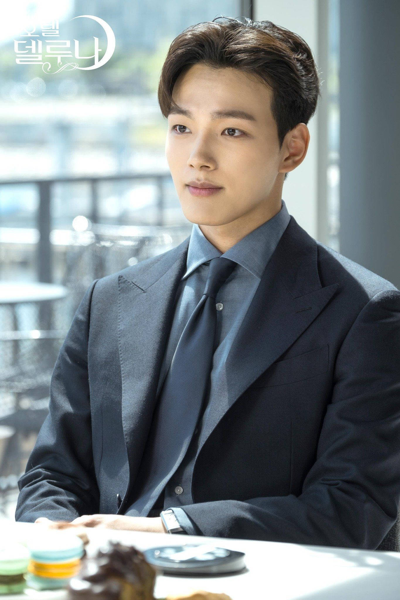 Yeo Jin Goo 為 TVN 拍攝劇照 壁紙 — 一位英俊的男子穿著俐落的深色西裝，坐在咖啡廳的桌子旁，桌上放著糕點，若有所思地望著明亮的窗外。