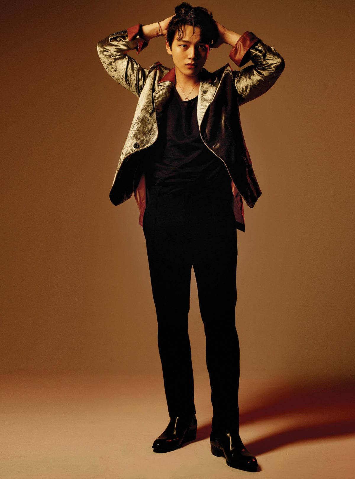 Yeo Jin Goo 銀紅色大衣 壁紙 — 一位身穿金屬外套和黑色服裝的年輕男子，在溫暖的棕色工作室背景前，自信地將雙手放在頭髮上擺姿勢。