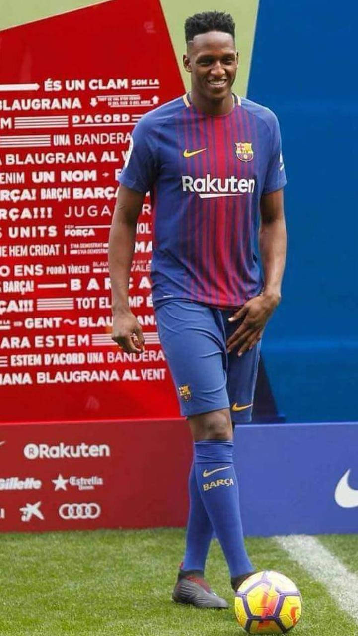 Yerry Mina 藍色球衣 壁紙 — 一位微笑的足球員身穿藍紅間條的FC巴塞隆拿球衣，站在綠色球場上，旁邊有一個色彩繽紛的足球。