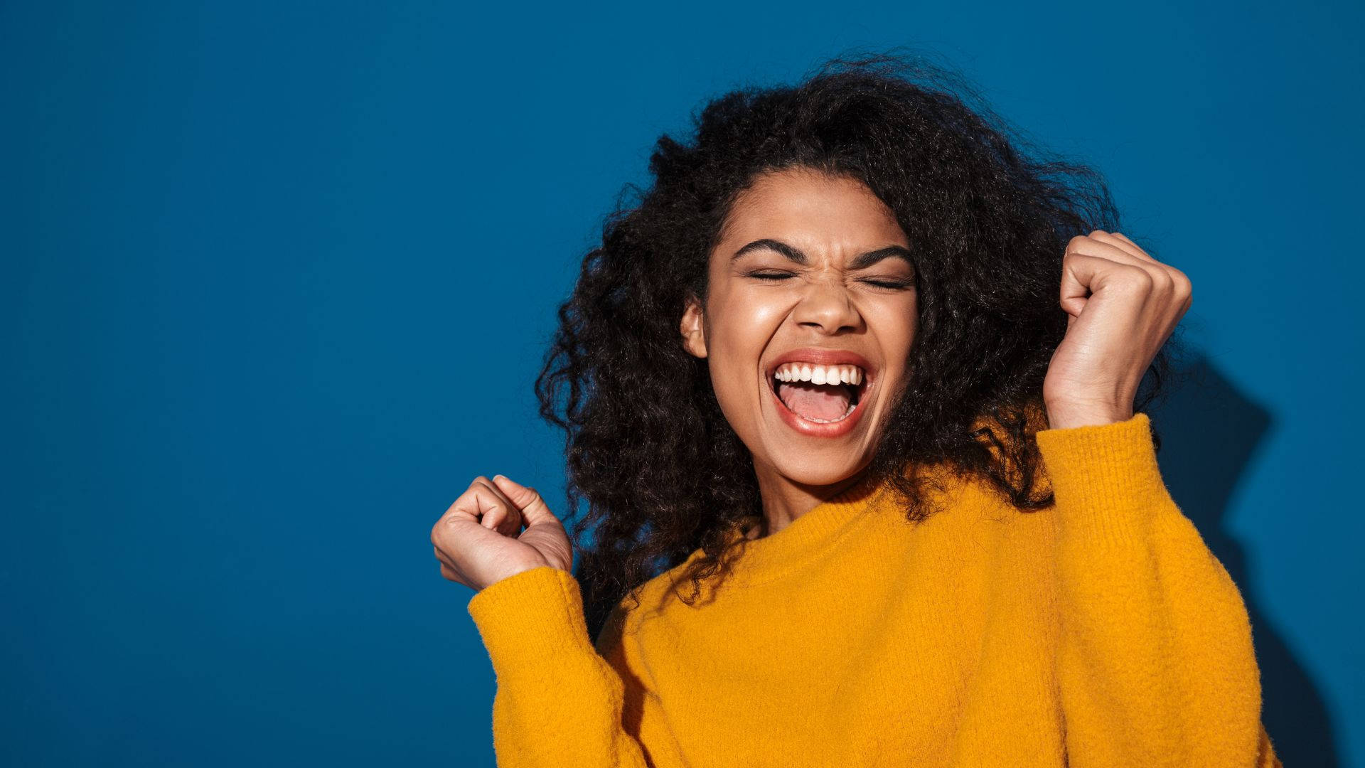 非洲女性大喊「Yey」 壁紙 — 一位快樂的捲髮女性穿著黃色毛衣，在純藍色背景前握緊拳頭慶祝。