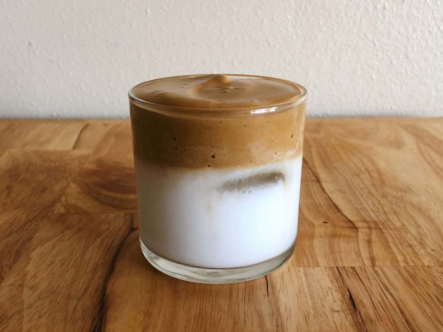  壁紙 — 一杯冰咖啡，帶有 creamy 白色牛奶和棕色咖啡層，放在溫暖的木桌上，背景是素色牆壁。