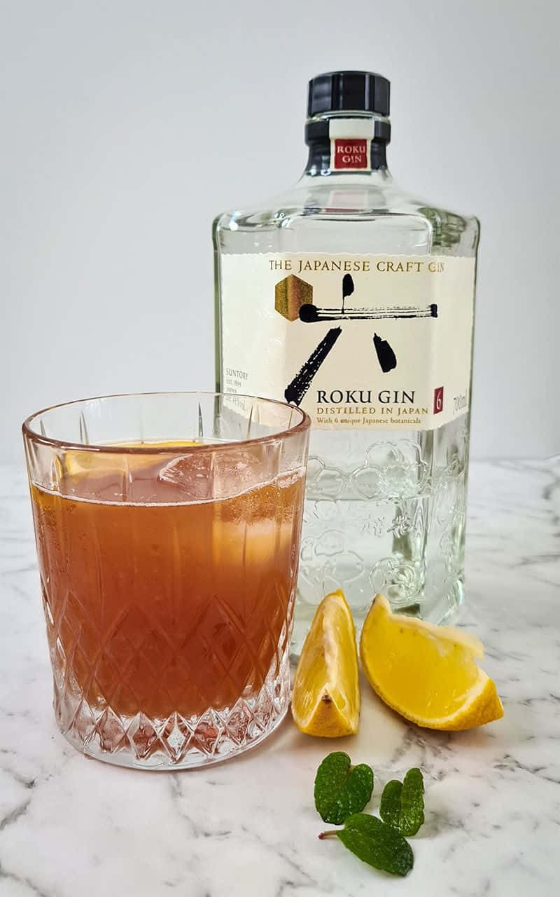  壁紙 — 一杯琥珀色氈酒雞尾酒加冰的晶瑩玻璃杯，旁邊放著一瓶 Roku Gin 和檸檬角，置於大理石表面上。