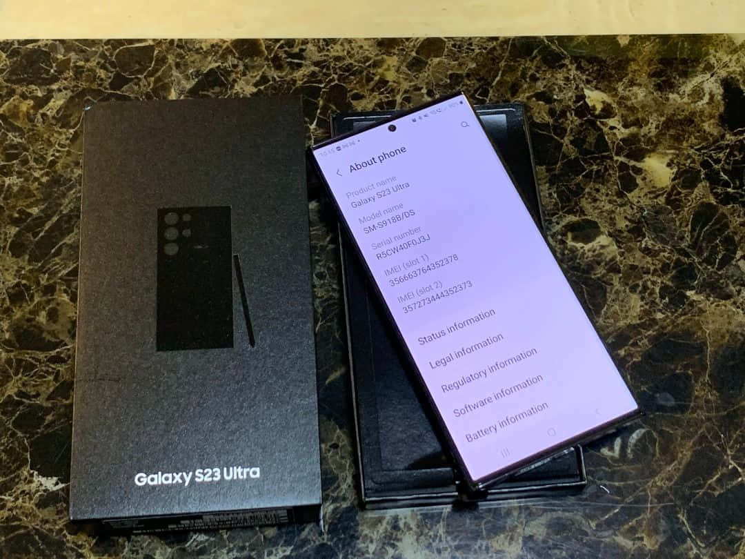Samsung S23 Ultra 桌布 壁紙 — 一部Samsung Galaxy S23 Ultra智能手機在深色大理石檯面上，於其黑色盒子旁邊顯示關於手機畫面。