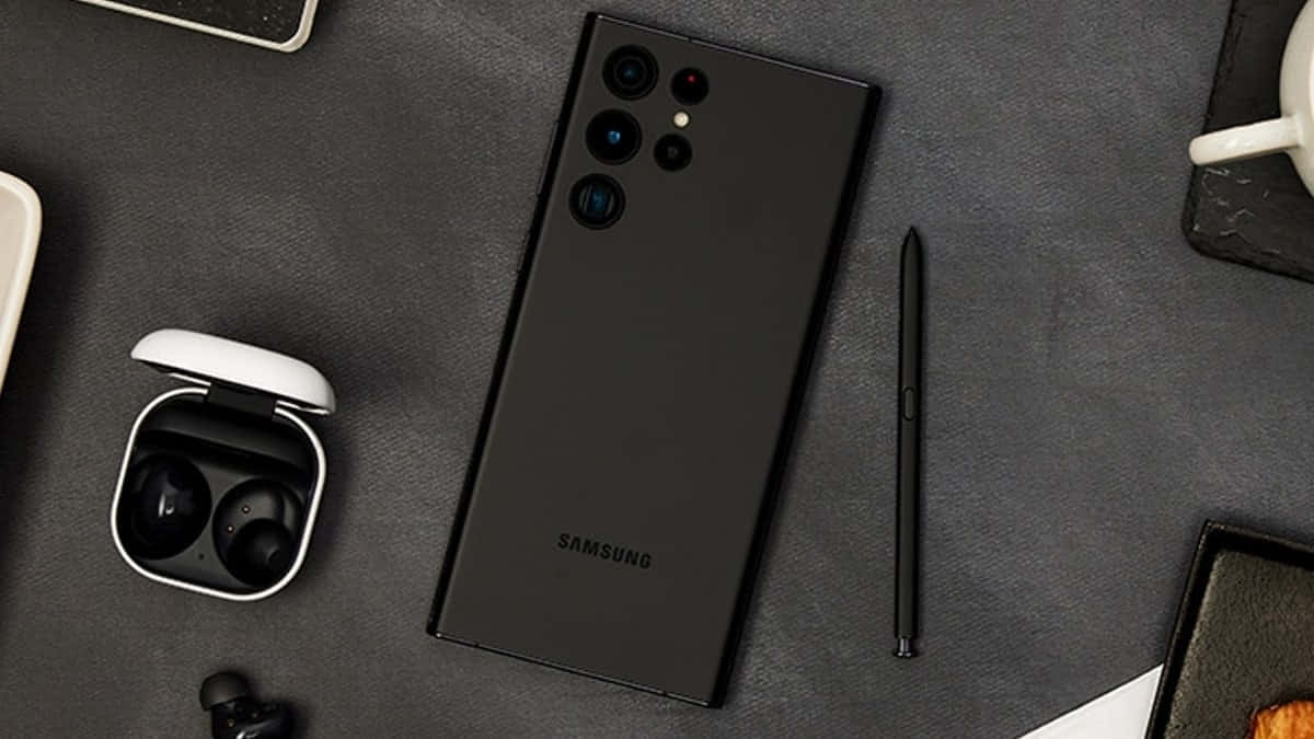 Samsung S23 Ultra 桌布 壁紙 — 一部時尚的黑色Samsung智能手機置於深灰色紋理表面中央，周圍有S Pen、耳機和一個白色咖啡杯。
