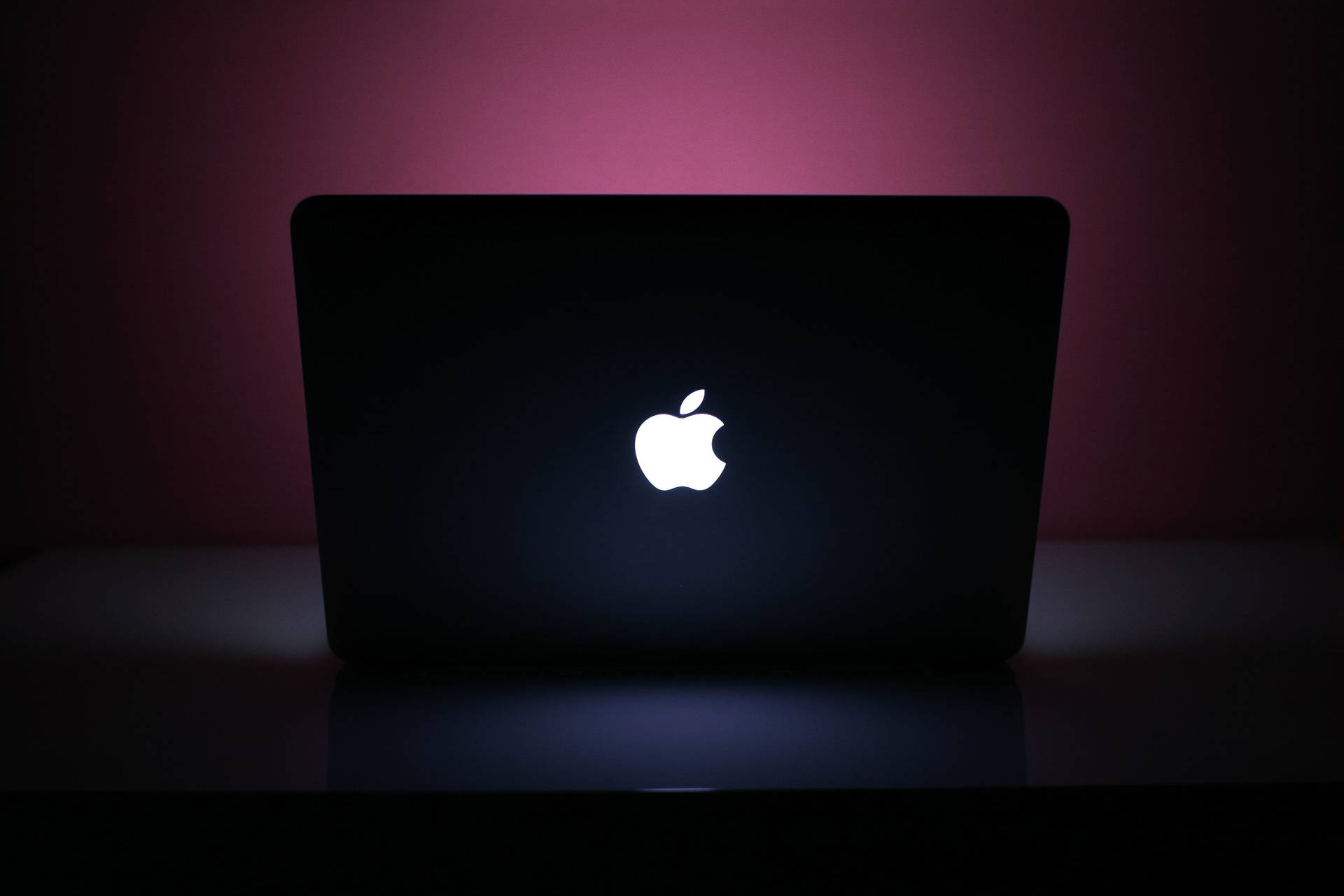 黑色手提電腦，帶有發光的Apple標誌，在粉紅色燈光下的深色表面上。