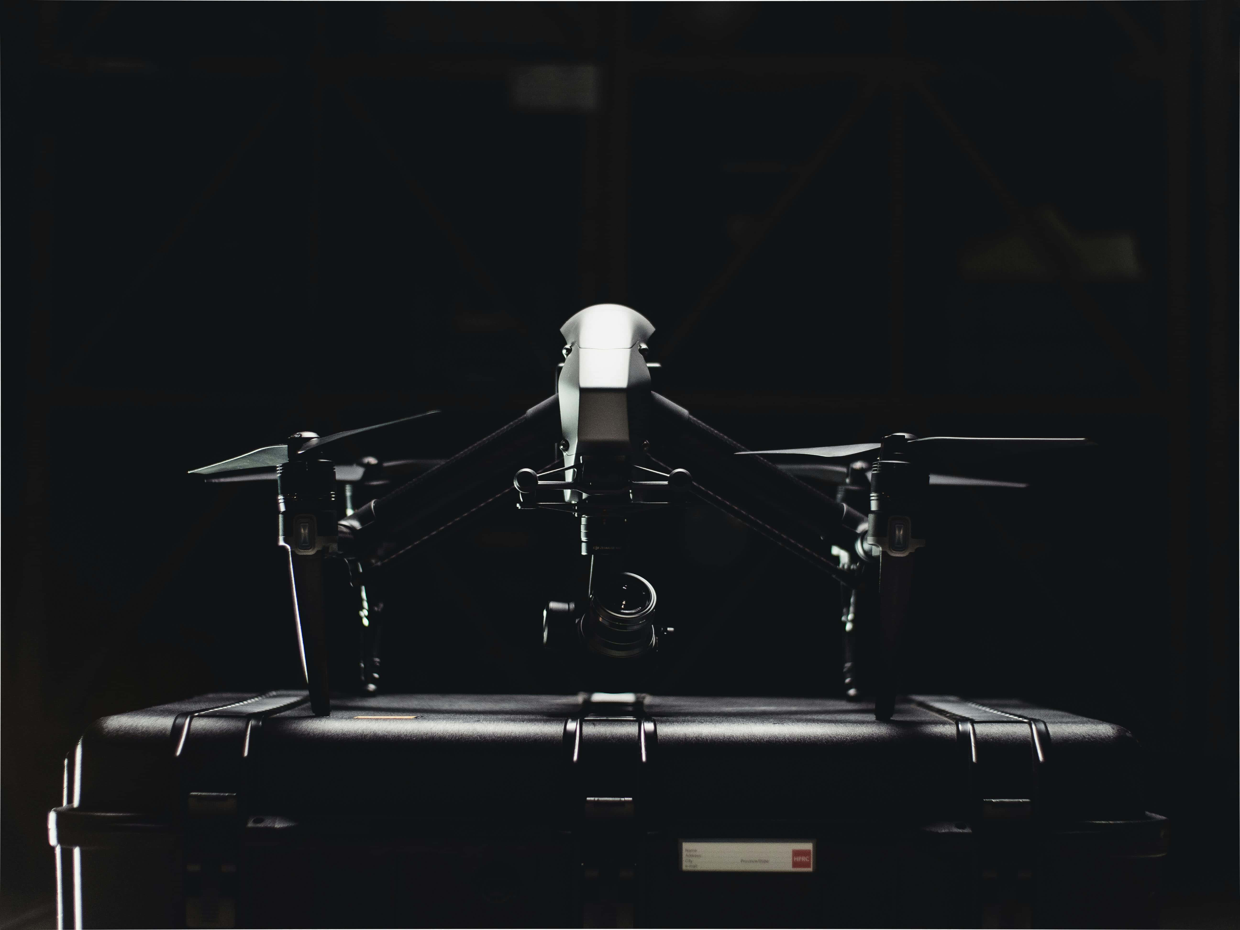 Drone Camera 桌布 壁紙 — 一部時尚的黑色無人機棲息在堅固的保護殼上，處於昏暗的工業空間，帶有戲劇性的背光效果。