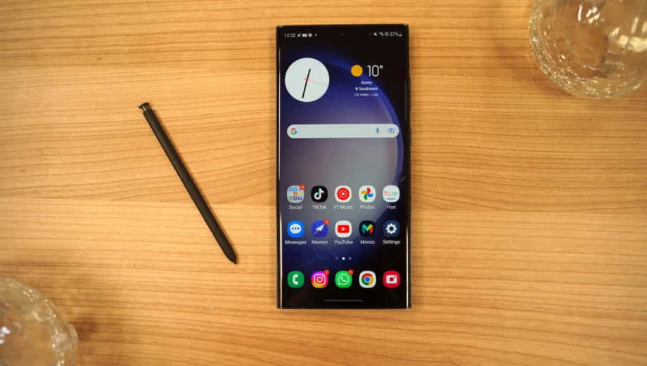 Samsung S23 Ultra 桌布 壁紙 — 一部時尚的黑色智能手機配備觸控筆，放在溫暖的木桌上，旁邊有玻璃器皿。