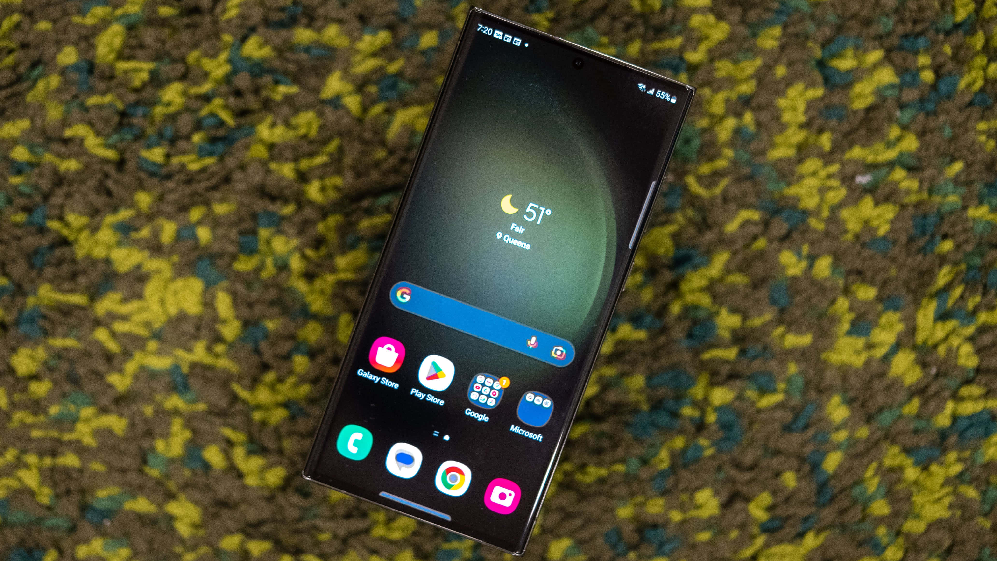 Samsung S23 Ultra 桌布 壁紙 — 一部時尚的黑色智能手機在質感黃綠色布料背景上顯示天氣小工具和應用程式圖示。