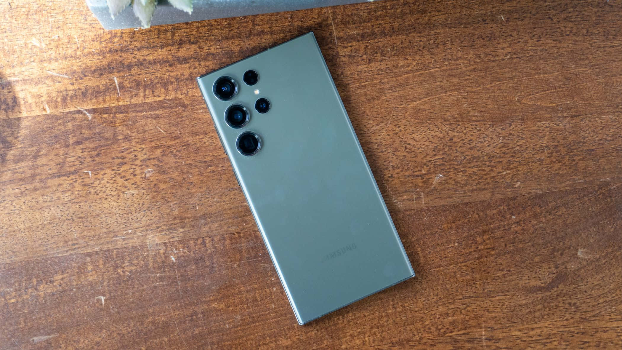 Samsung S23 Ultra 桌布 壁紙 — 一部時尚的鼠尾草綠色Samsung智能手機以對角線方式放置在溫暖有紋理的木質表面上，伴隨著柔和自然光。