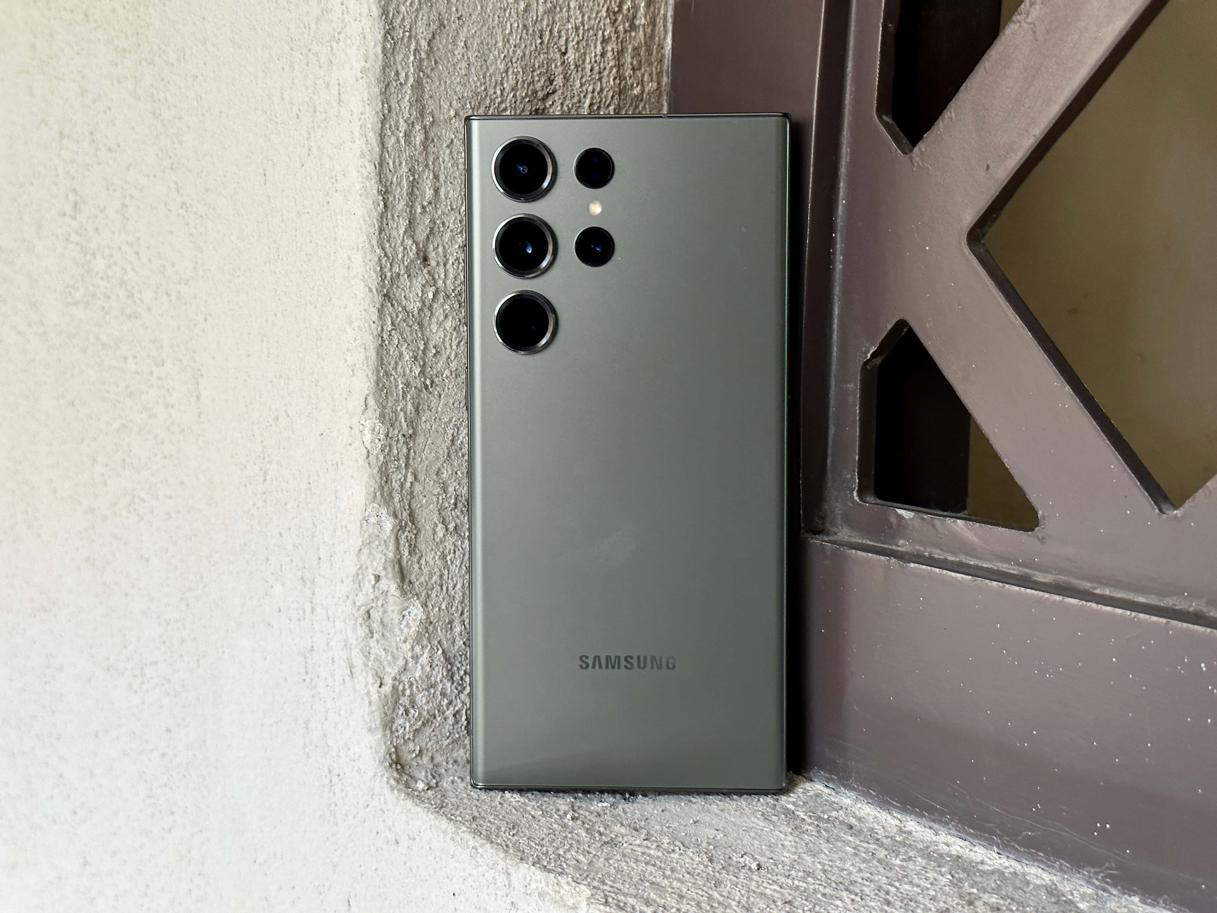 Samsung S23 Ultra 桌布 壁紙 — 一部時尚的啞光綠色Samsung智能手機靠在一面有紋理的牆上，旁邊是一扇幾何金屬門。