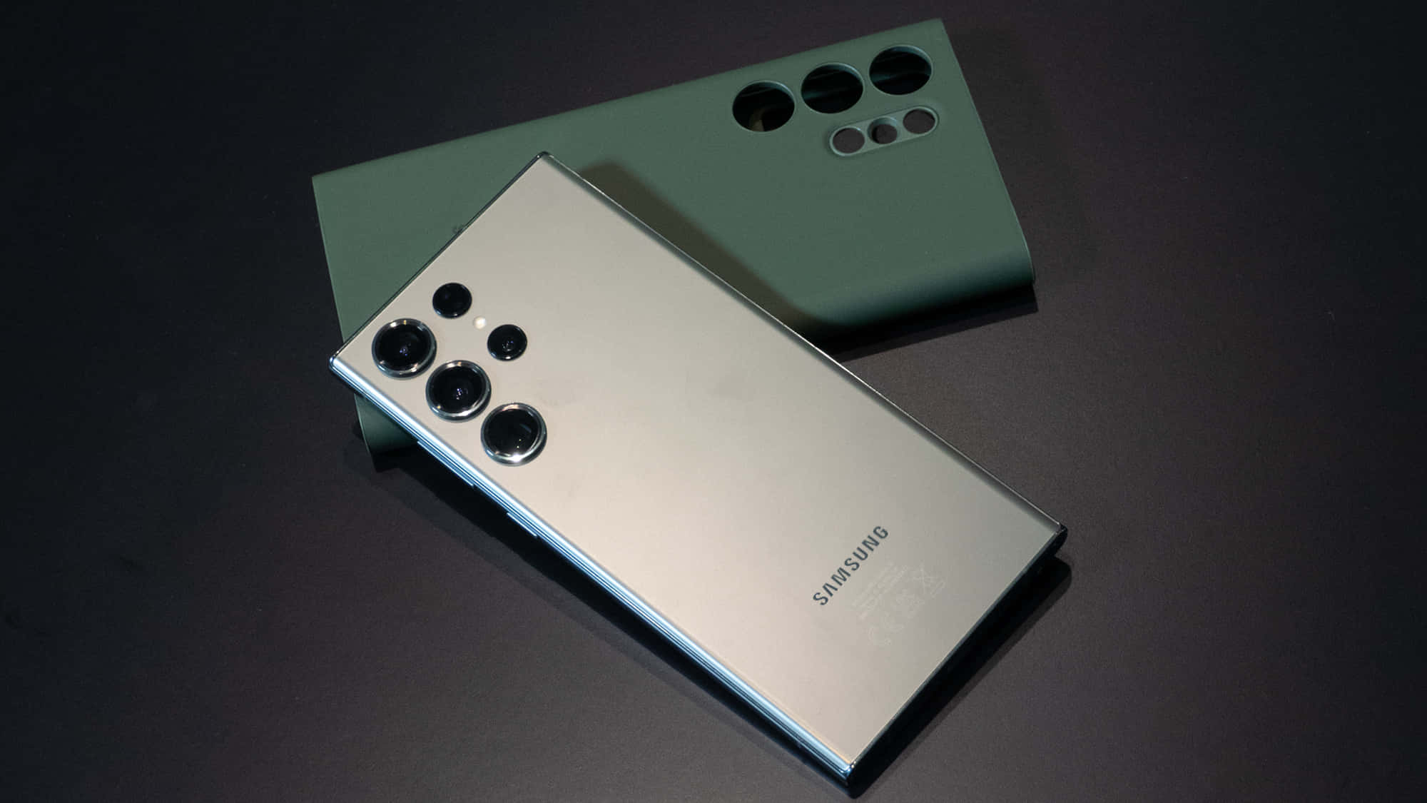 Samsung S23 Ultra 桌布 壁紙 — 一部時尚的銀色Samsung智能手機斜放在啞光綠色手機殼上，置於深色表面。