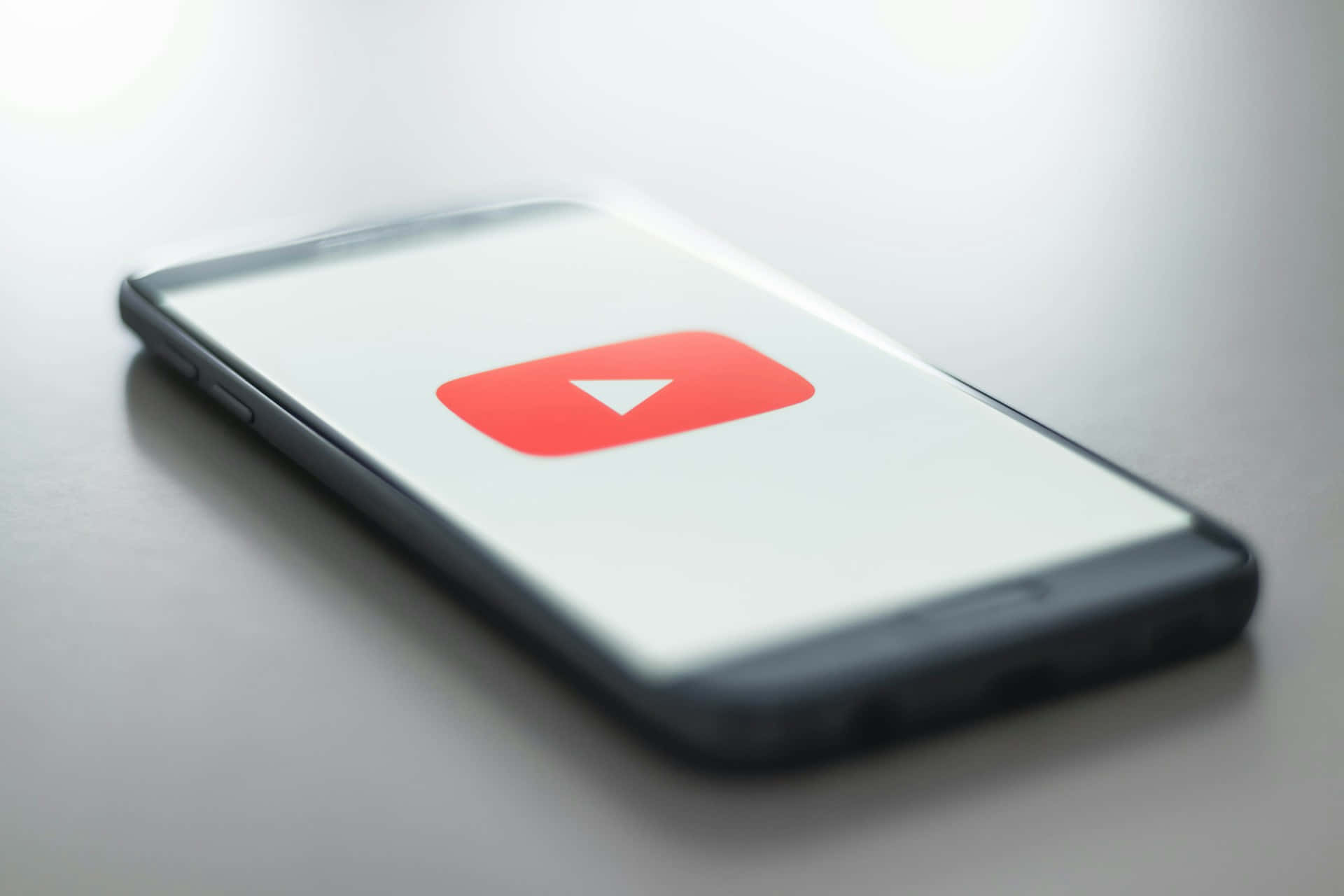  壁紙 — 一部時尚的智能手機在極簡白色屏幕上顯示標誌性的紅色 YouTube 播放按鈕標誌。