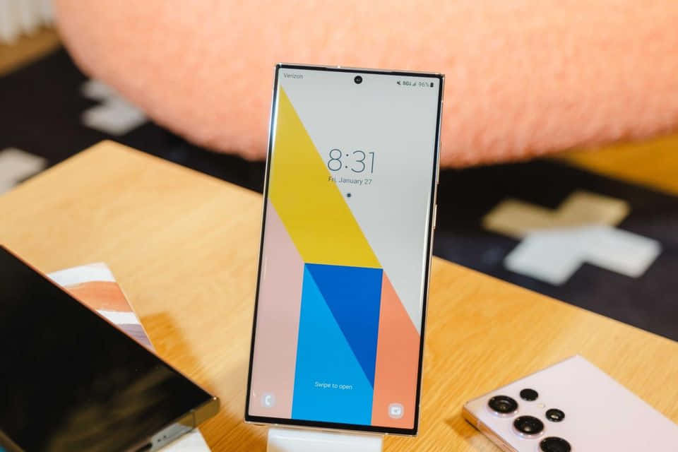 Samsung S23 Ultra 桌布 壁紙 — 一部時尚智能手機在木桌上顯示色彩繽紛的幾何圖案壁紙，背景有一個柔軟的粉紅色靠墊。