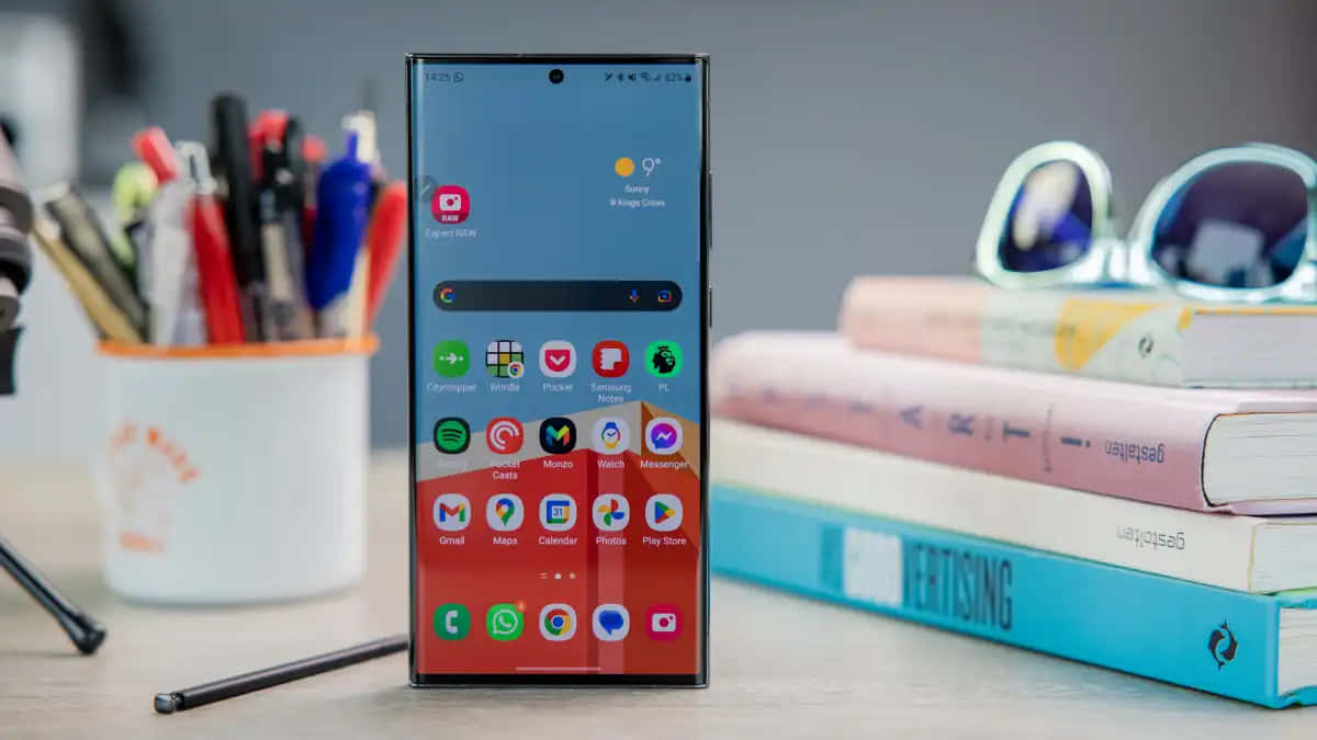 Samsung S23 Ultra 桌布 壁紙 — 一部時尚智能手機在木桌上展示充滿活力的主屏幕，旁邊有書籍、太陽眼鏡和筆筒。