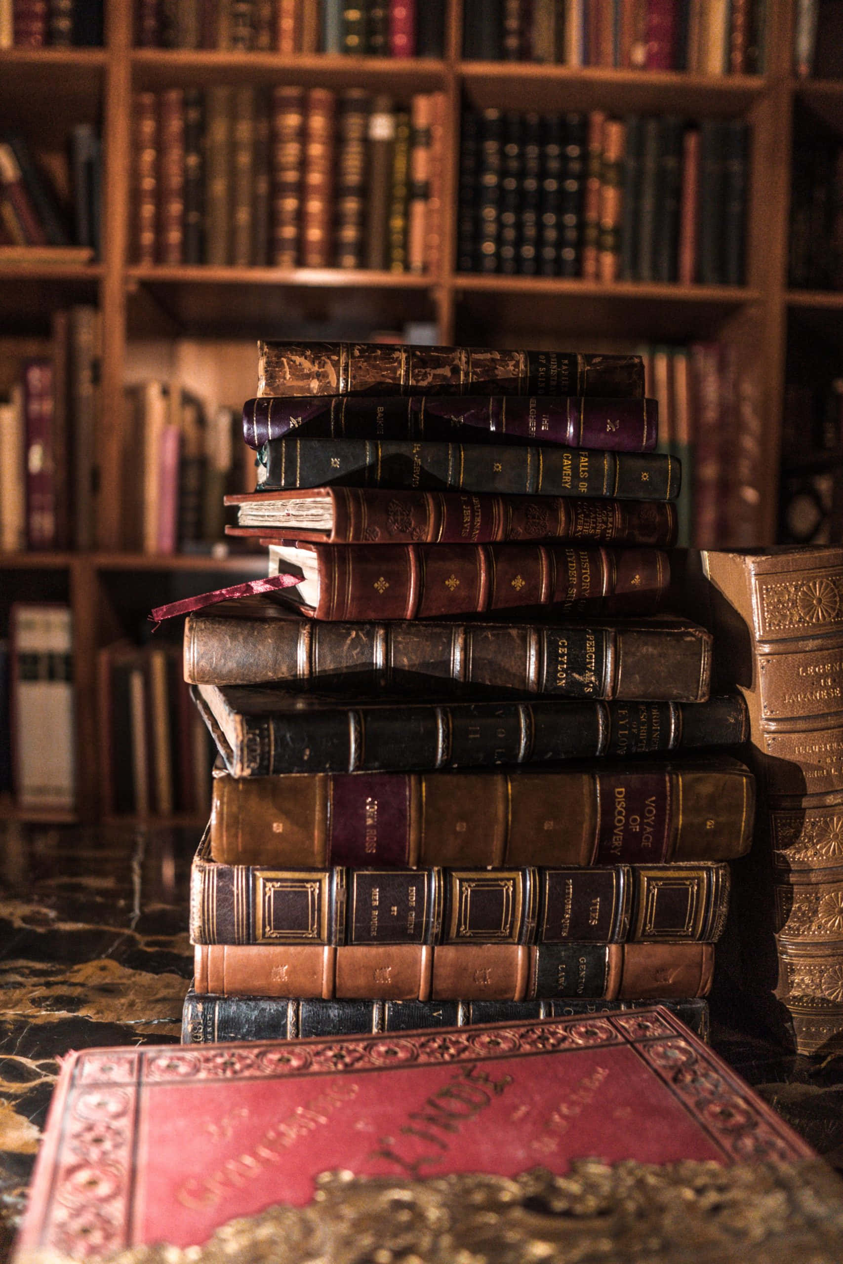 Dark Book Aesthetic 桌布 壁紙 — 一堆復古皮革裝訂的書籍高高堆疊在大理石桌上，旁邊的書架上放滿了陳舊的書卷，書房內燈光溫暖。