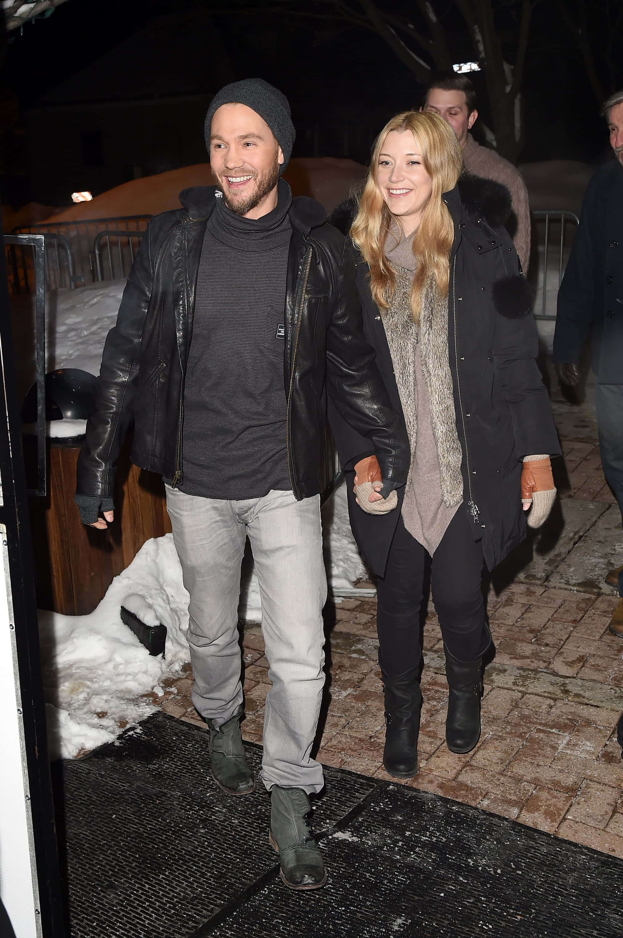 Sarah Roemer 桌布 壁紙 — 一對微笑的情侶手牽手走過積雪的夜晚街道，穿著冬季外套和手套。