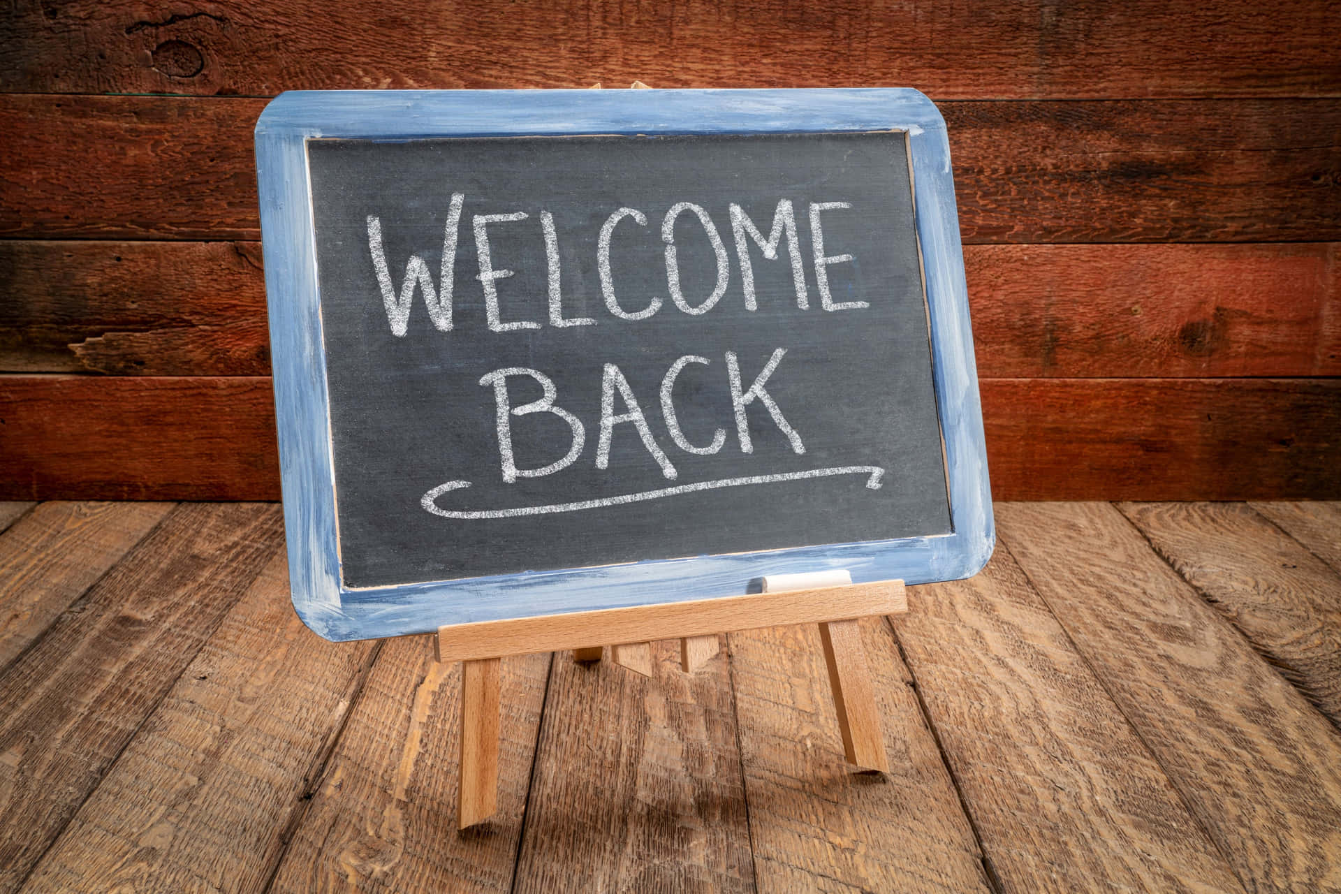  壁紙 — 一塊寫著welcome back的黑板標誌放在木製畫架上，背景是質樸的木牆。
