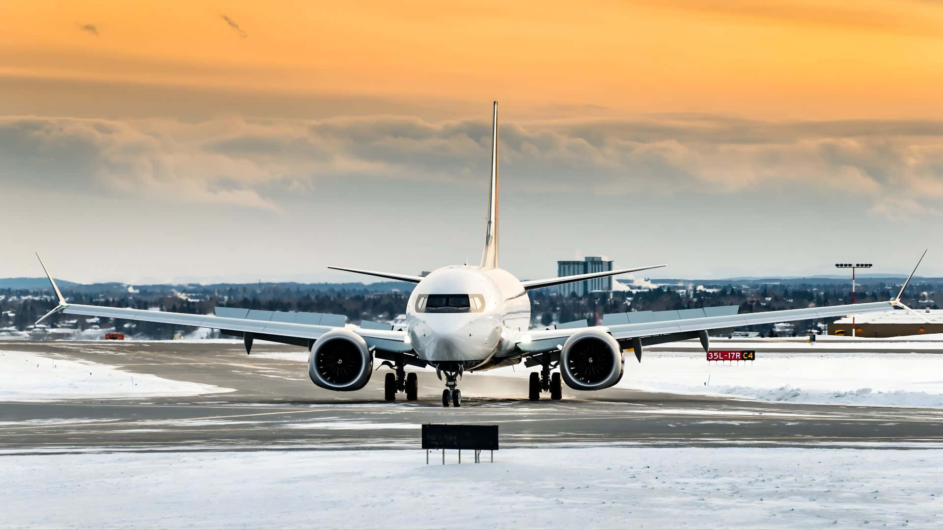  壁紙 — 一架白色客機停在黃昏時分積雪的跑道上，上空有橙色雲朵，遠處有城市建築。
