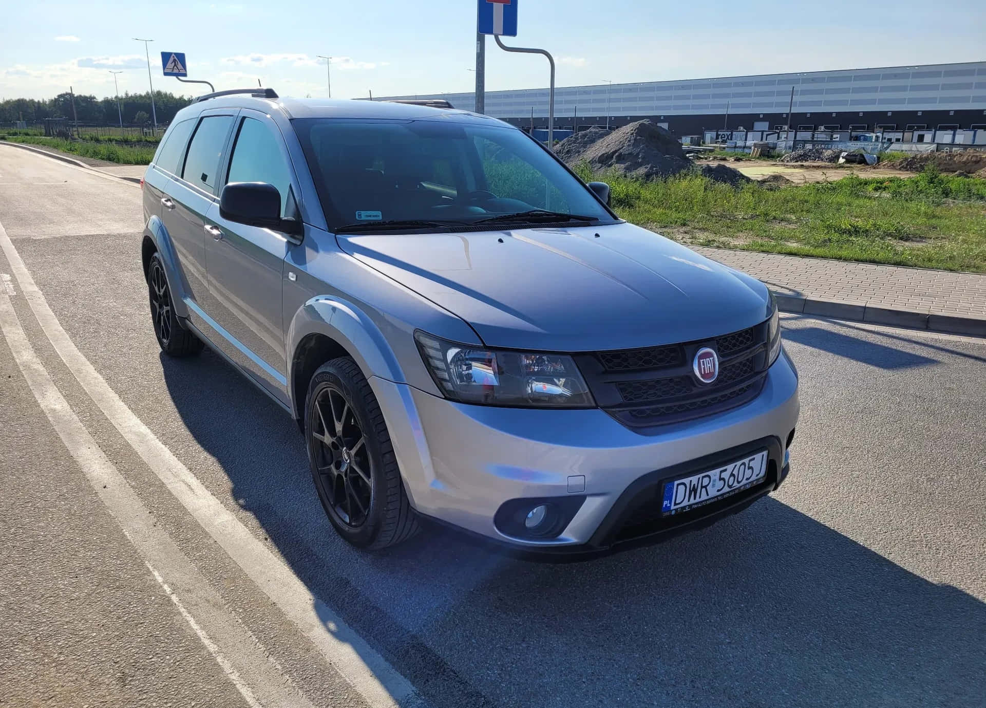  壁紙 — 一架銀色Fiat SUV停泊在陽光明媚的路邊，配有黑色輪圈，背景是現代工業建築。