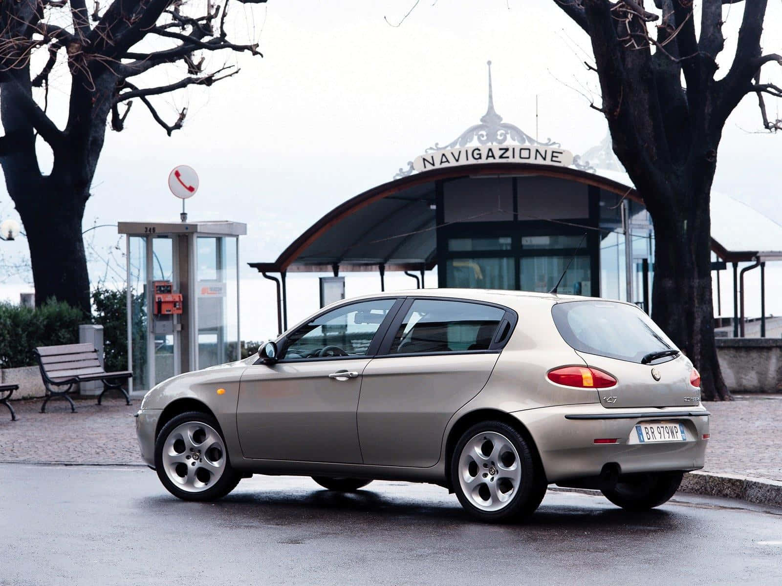Alfa Romeo 147 桌布 壁紙 — 一架銀色愛快羅密歐147停泊在濕漉漉的路面上，旁邊有電話亭和一個「Navigazione」候車亭，天空多雲。
