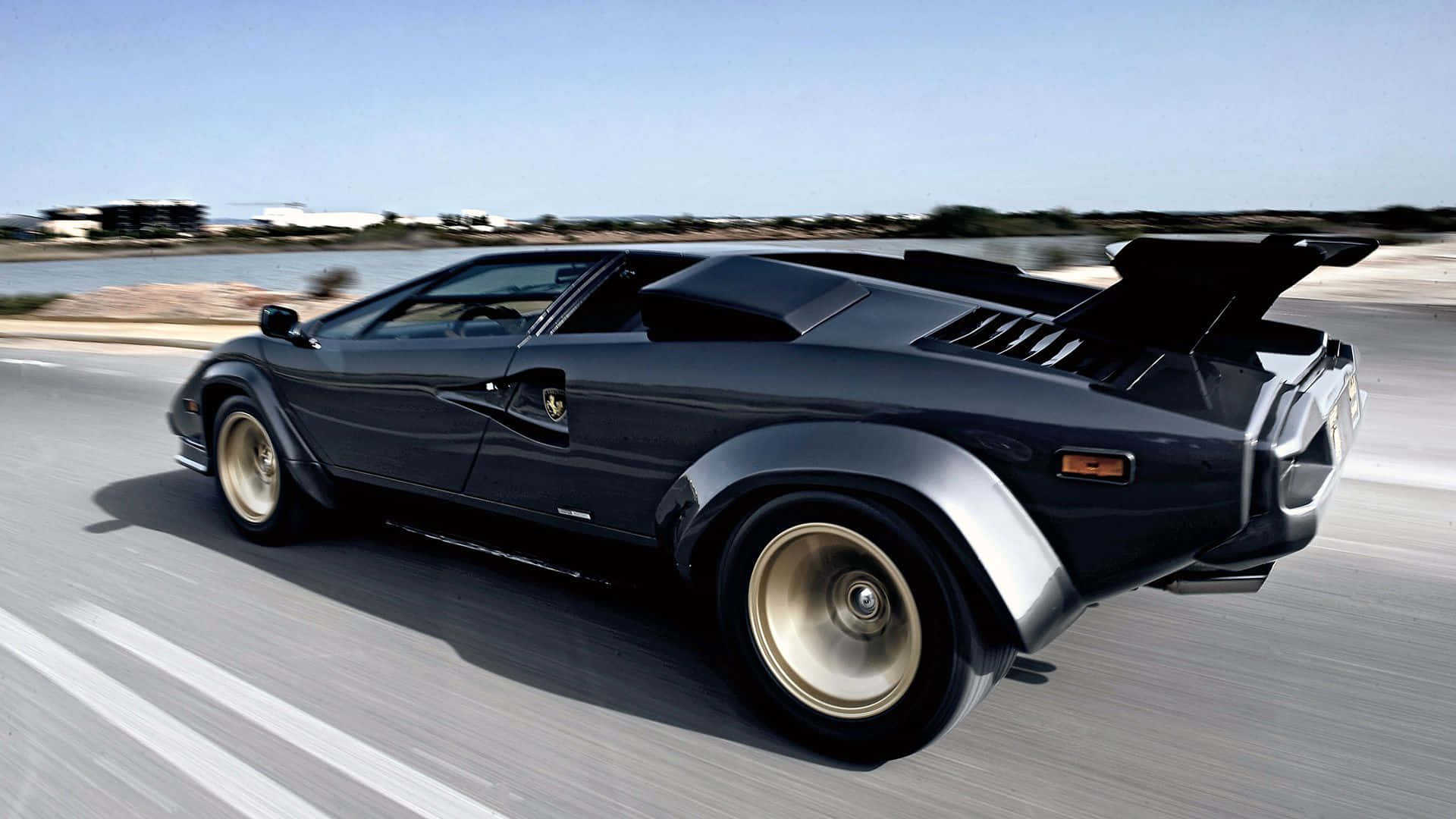 Lamborghini Countach 桌布 壁紙 — 一架時尚的黑色林寶堅尼Countach在晴朗藍天下沿著海岸高速公路飛馳。