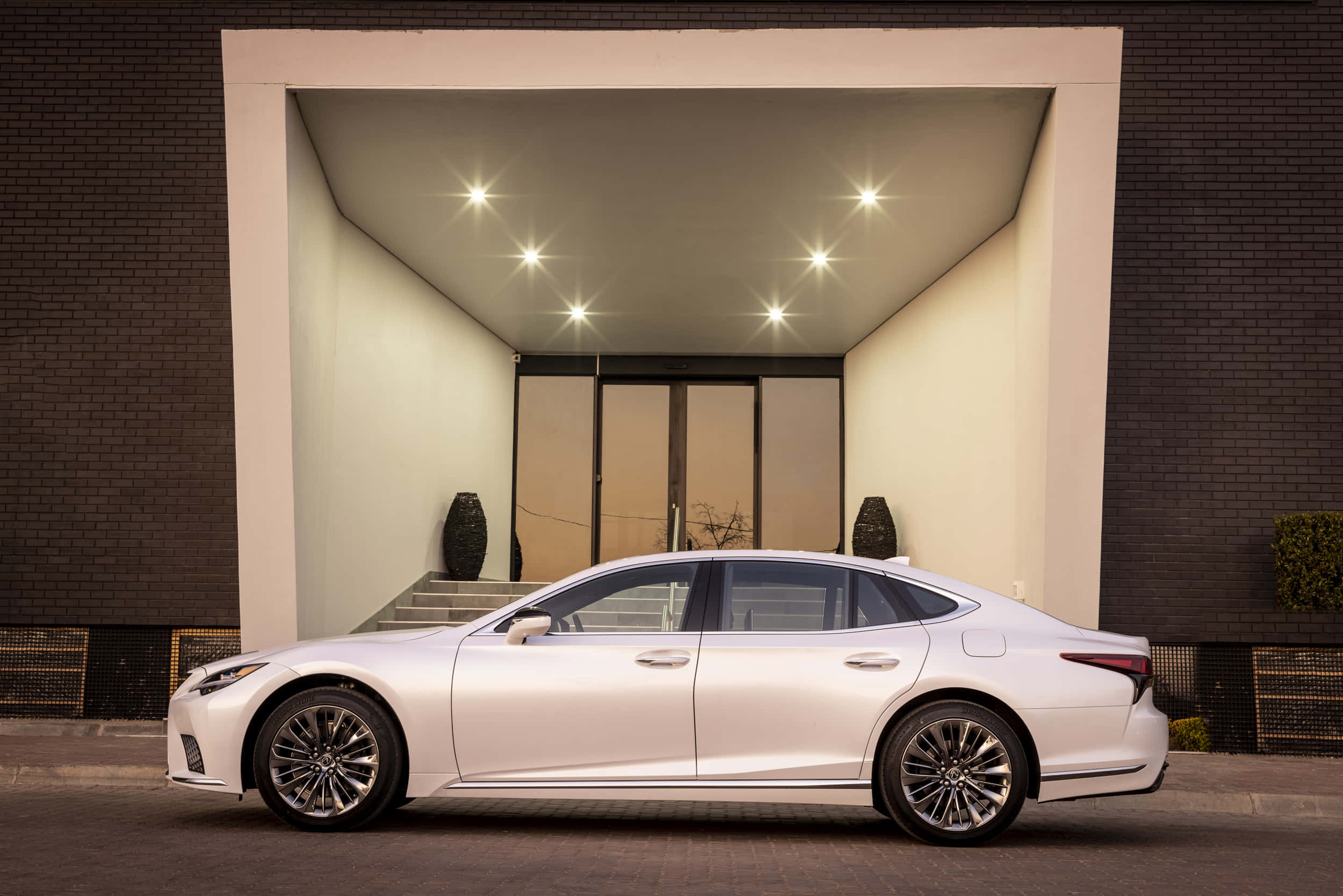 Lexus Ls 500 桌布 壁紙 — 一架時尚的白色豪華轎車停泊在現代建築風格的門廊下，配有嵌入式燈光和深色磚牆。