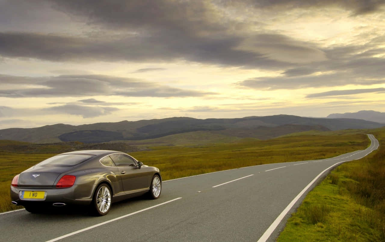 Bentley Continental Gt 桌布 壁紙 — 一架時尚的灰色賓利雙門跑車沿著蜿蜒的鄉村道路行駛，穿過起伏的綠色山丘，天空戲劇性地多雲。