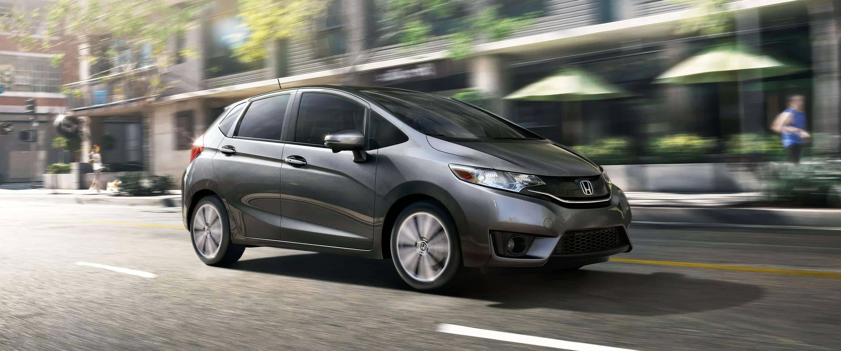 Honda Fit 桌布 壁紙 — 一架時尚的灰色本田飛度在陽光明媚的城市街道上飛馳，背景建築物和綠色遮陽篷呈現動態模糊效果。