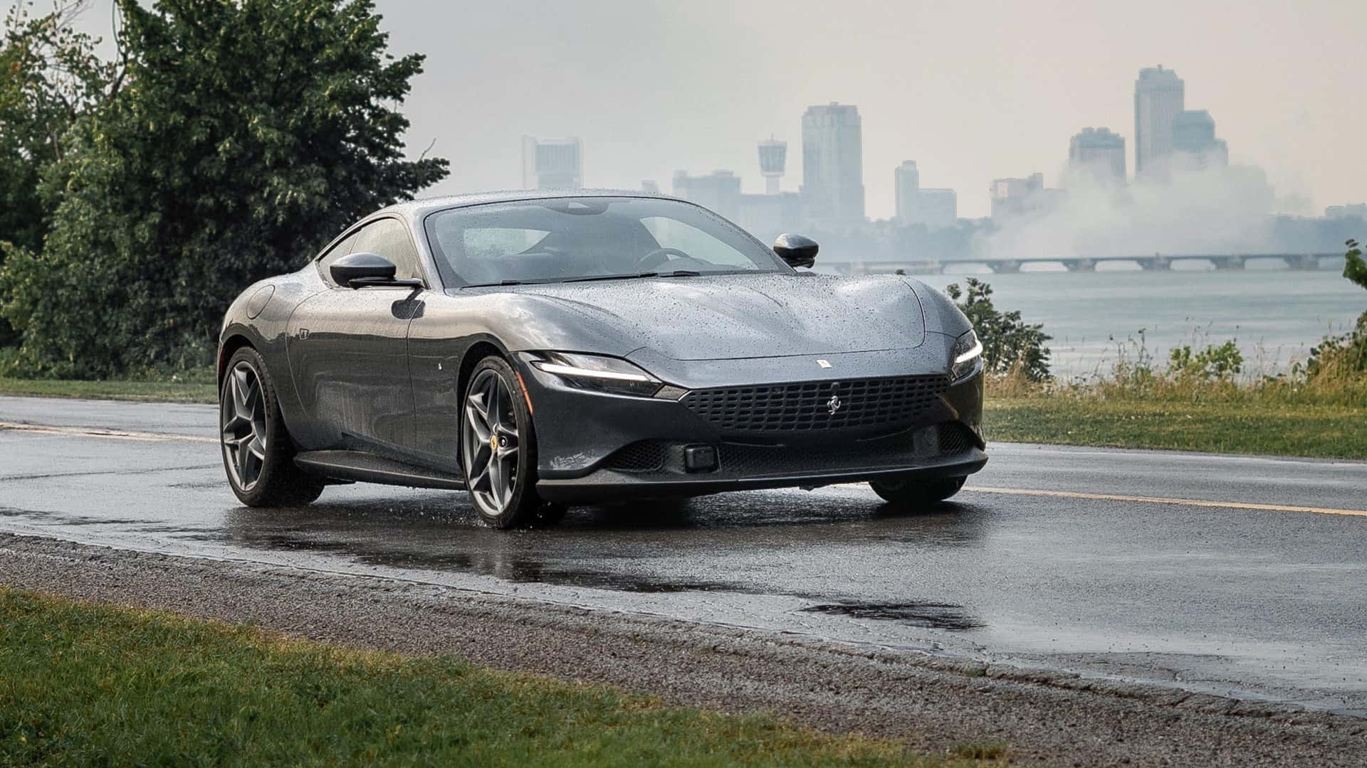 Ferrari Roma 桌布 壁紙 — 一架時尚的灰色法拉利在潮濕的道路上行駛，背景是霧濛濛的城市天際線和河流。