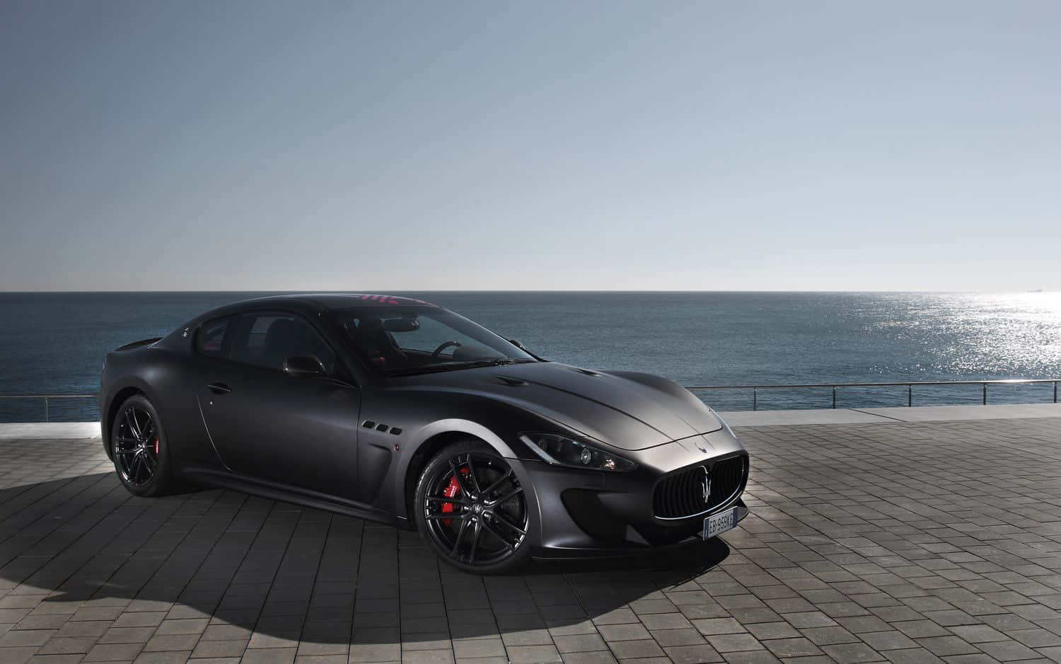 Maserati Granturismo 桌布 壁紙 — 一架時尚的啞光灰色瑪莎拉蒂跑車停泊在瓷磚鋪設的海濱長廊上，海平線在晴朗天空下清晰可見。