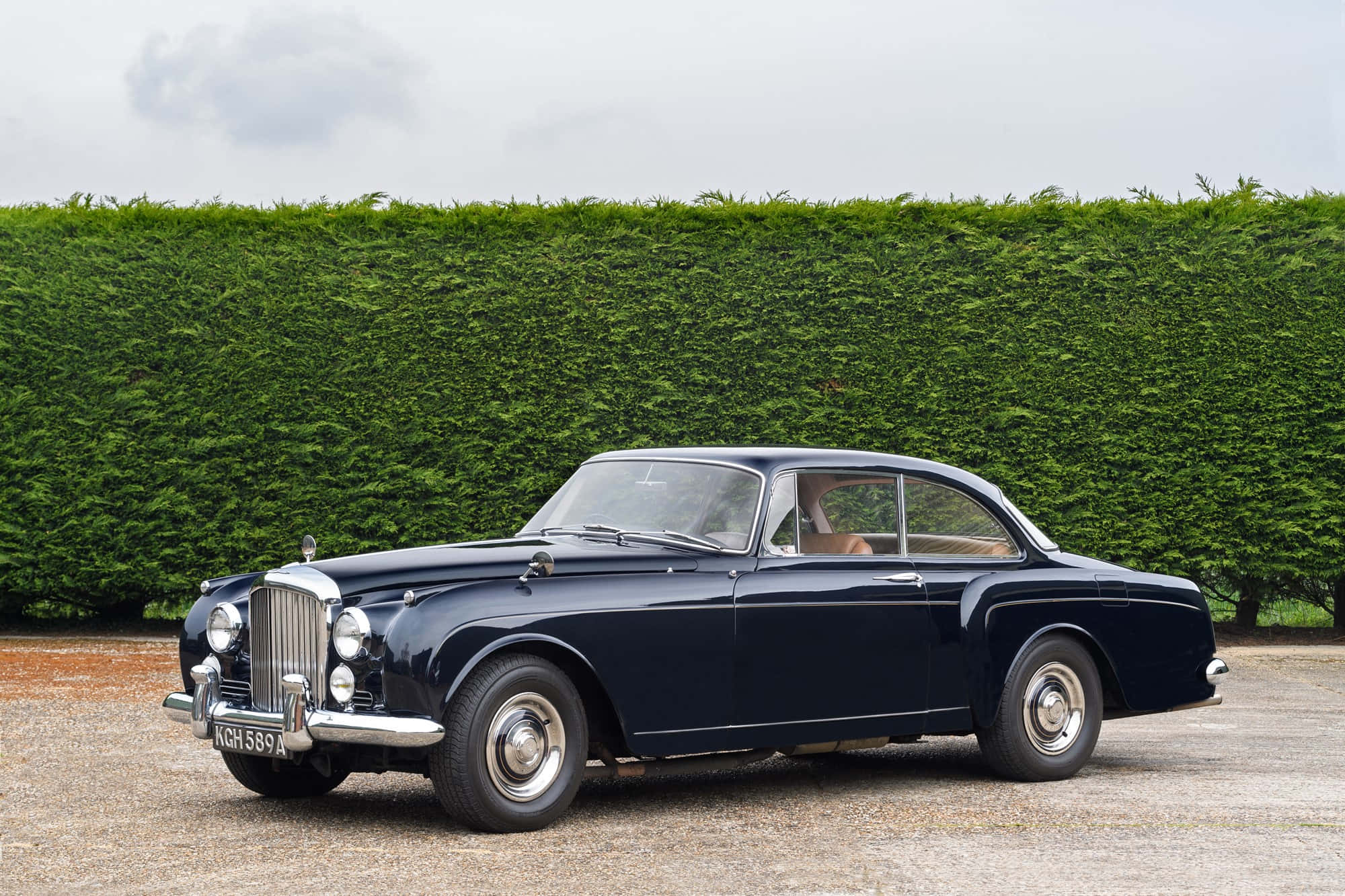 Bentley S2 桌布 壁紙 — 一架時尚的深藍色經典汽車，帶有鍍鉻裝飾，停泊在碎石路上，前面是高大的綠色樹籬，天空多雲。